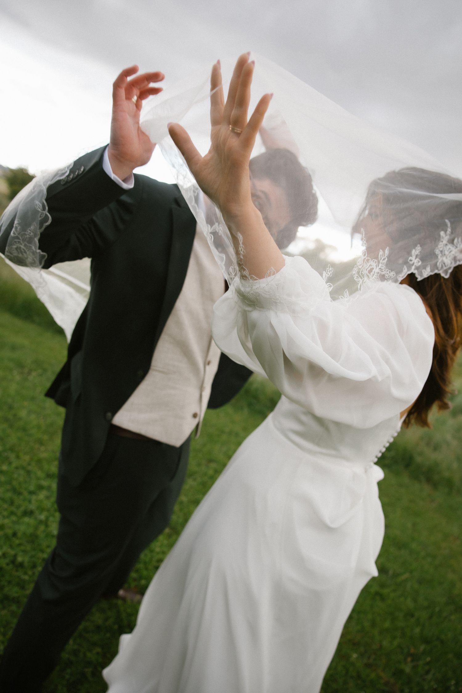 Bride and groom playfully interact under a billowing lace veil in an outdoor setting.