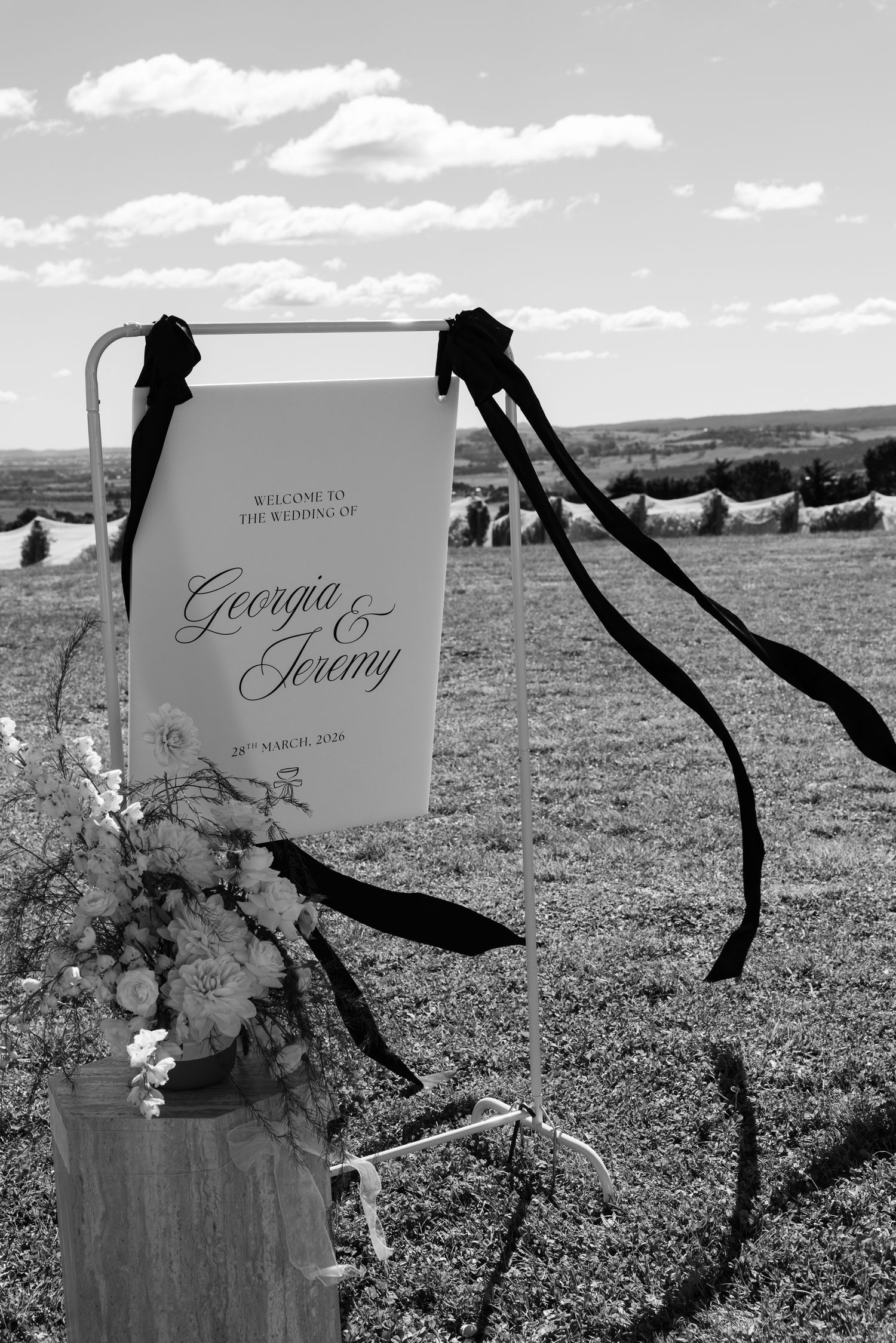 Black and white wedding welcome sign for Georgia and Jeremy displayed in sunny countryside field.