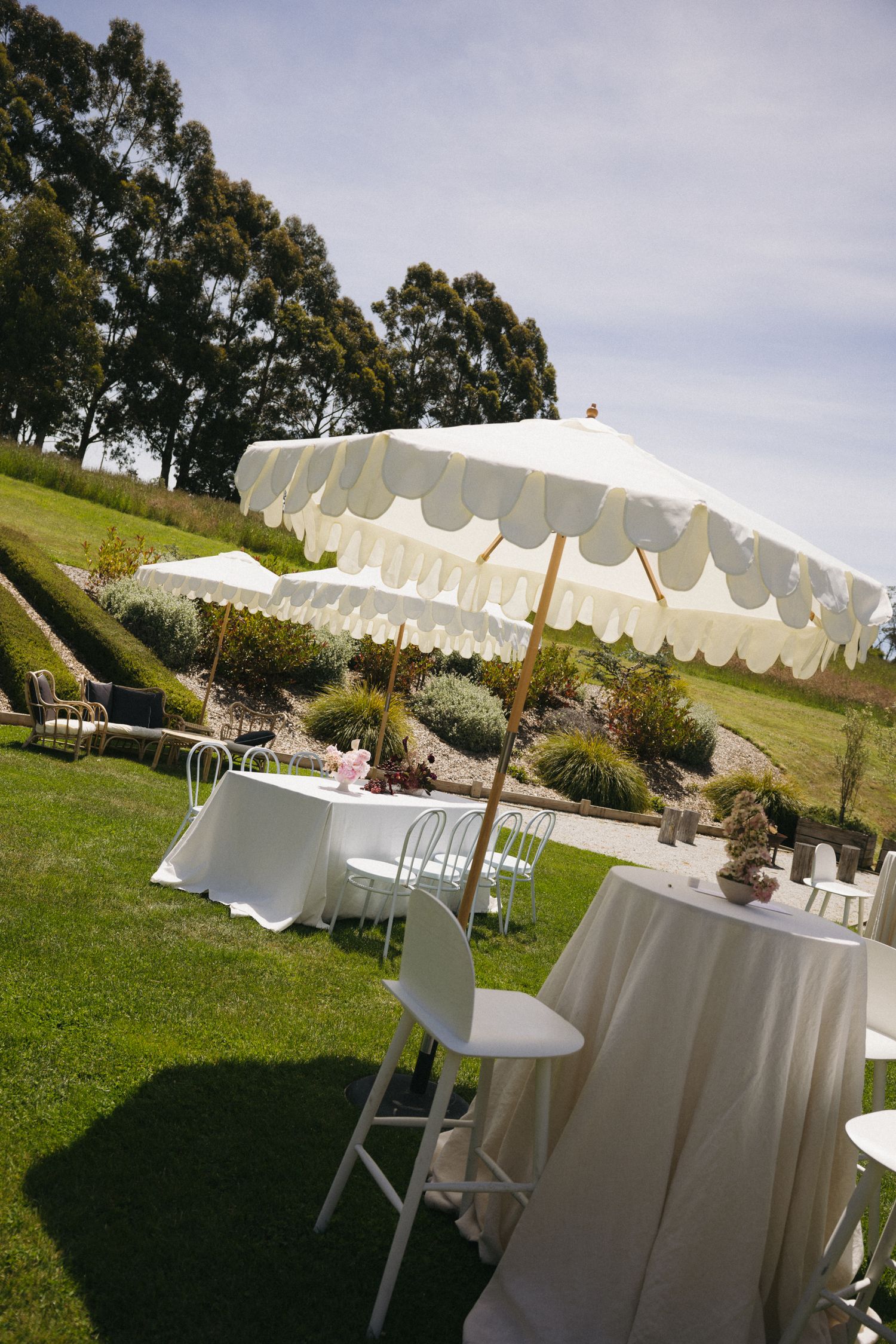 Elegant outdoor wedding reception with white umbrella tables and chairs on a lush green lawn.