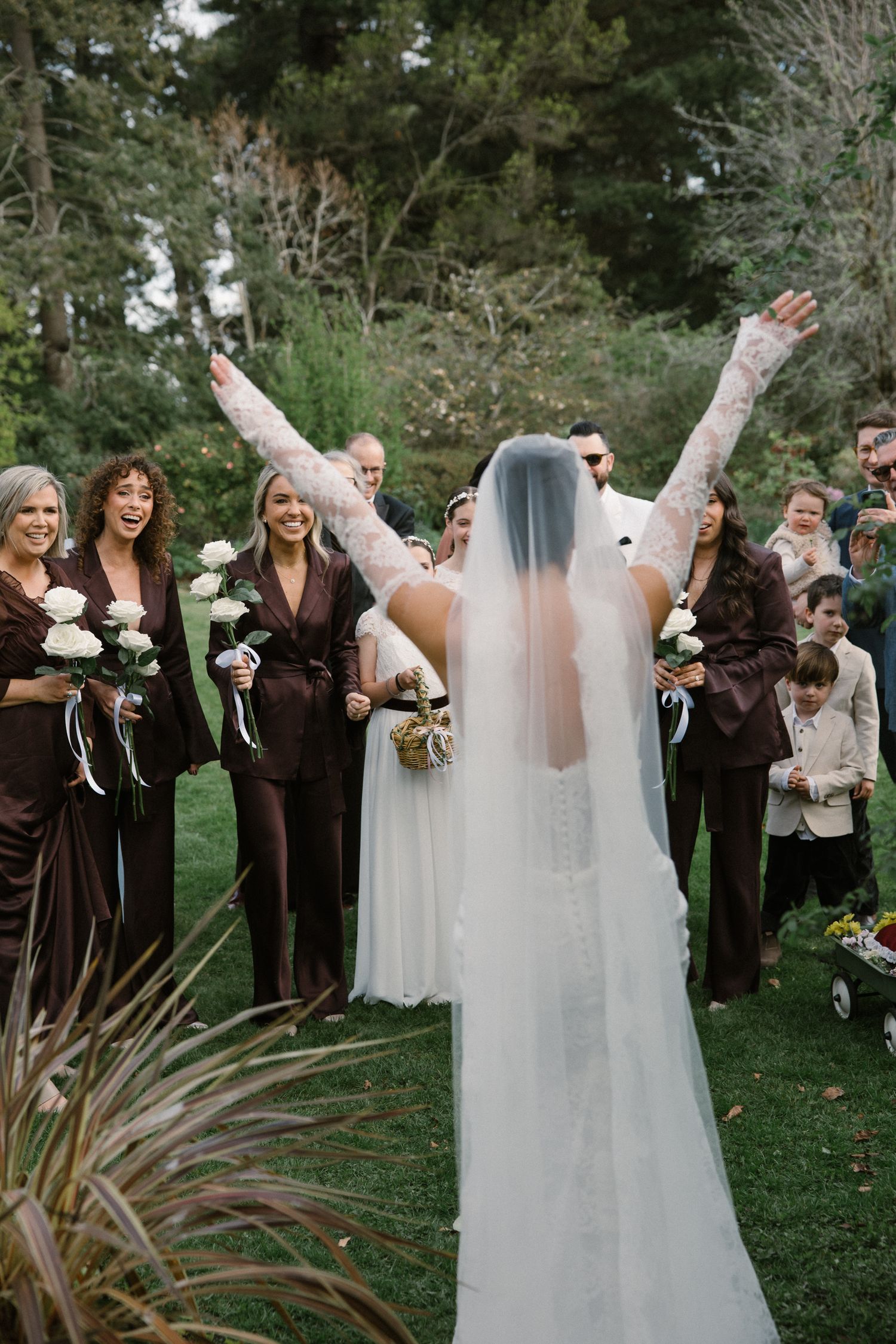 Bride in lace gown raises arms joyfully as bridesmaids in brown dresses celebrate outdoors.