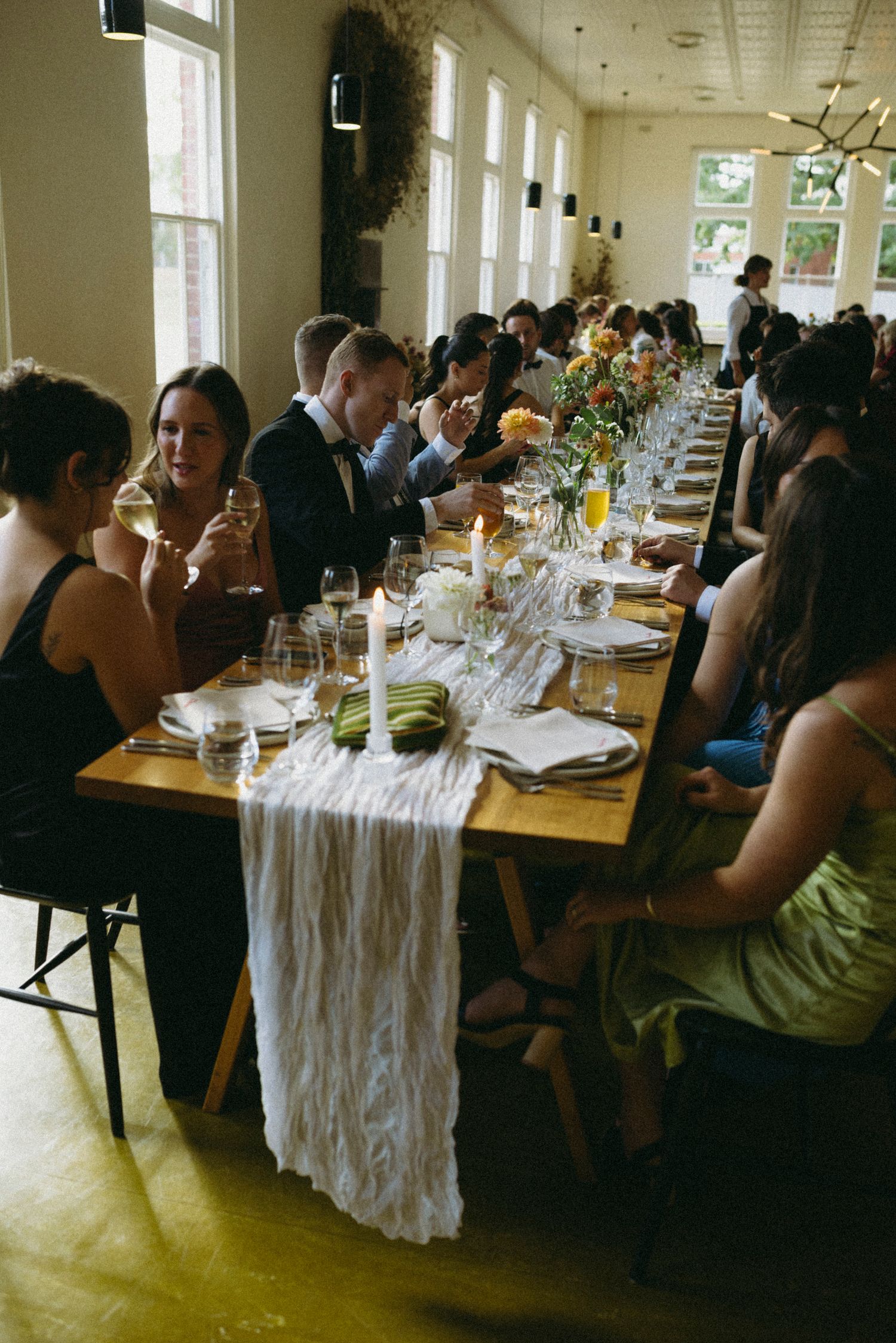 Elegant wedding reception with guests seated at long wooden tables adorned with white runners and floral centerpieces.