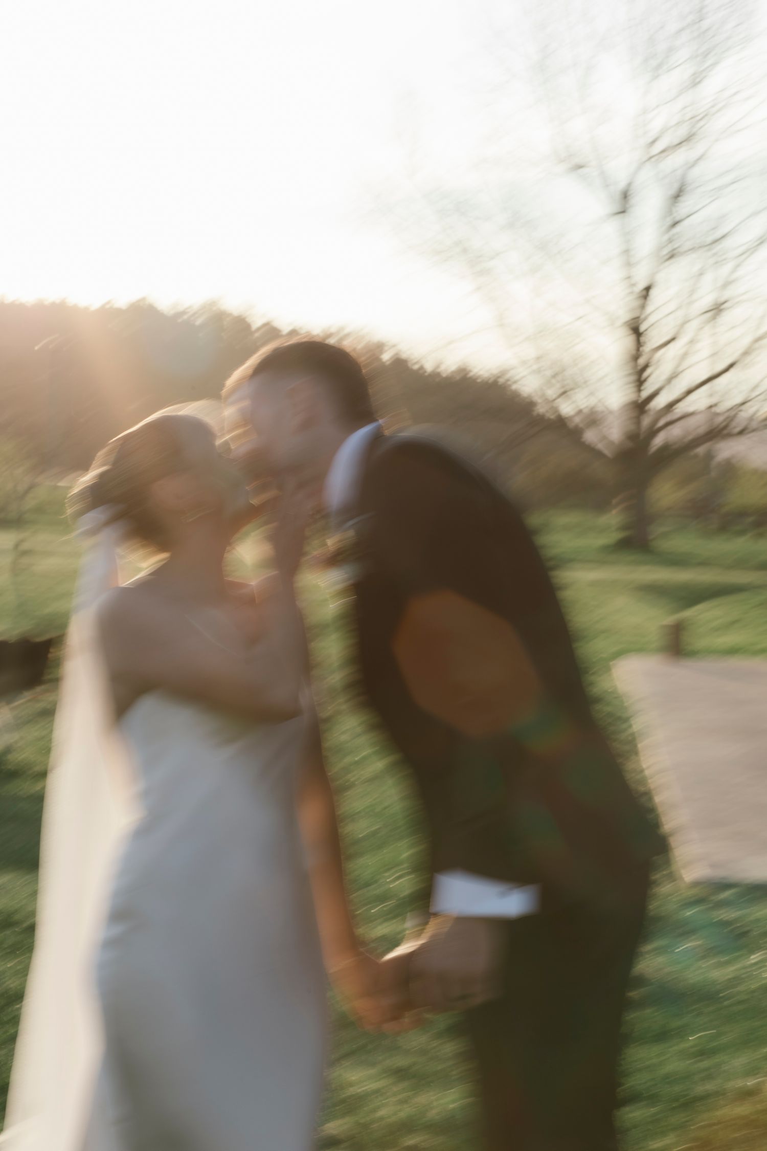 Artistic motion-blurred photo of bride and groom kissing at golden hour in an outdoor setting.