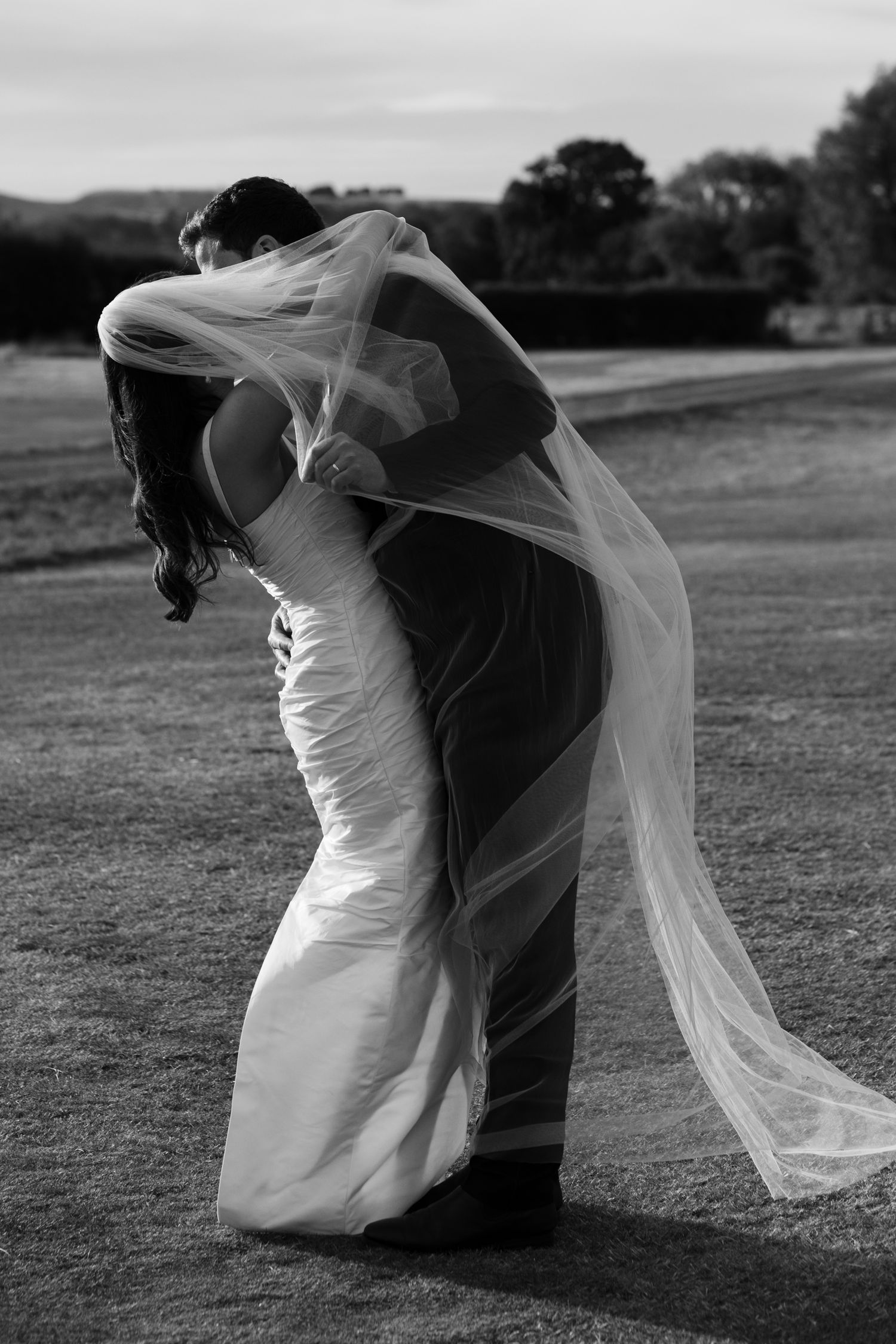 Black and white photo of bride and groom sharing romantic dip kiss with flowing veil.