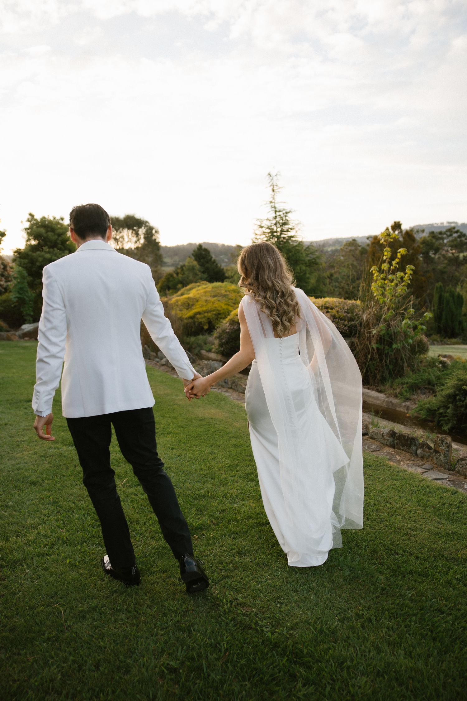 Bride and groom holding hands walking through lush green garden at sunset in elegant wedding attire.