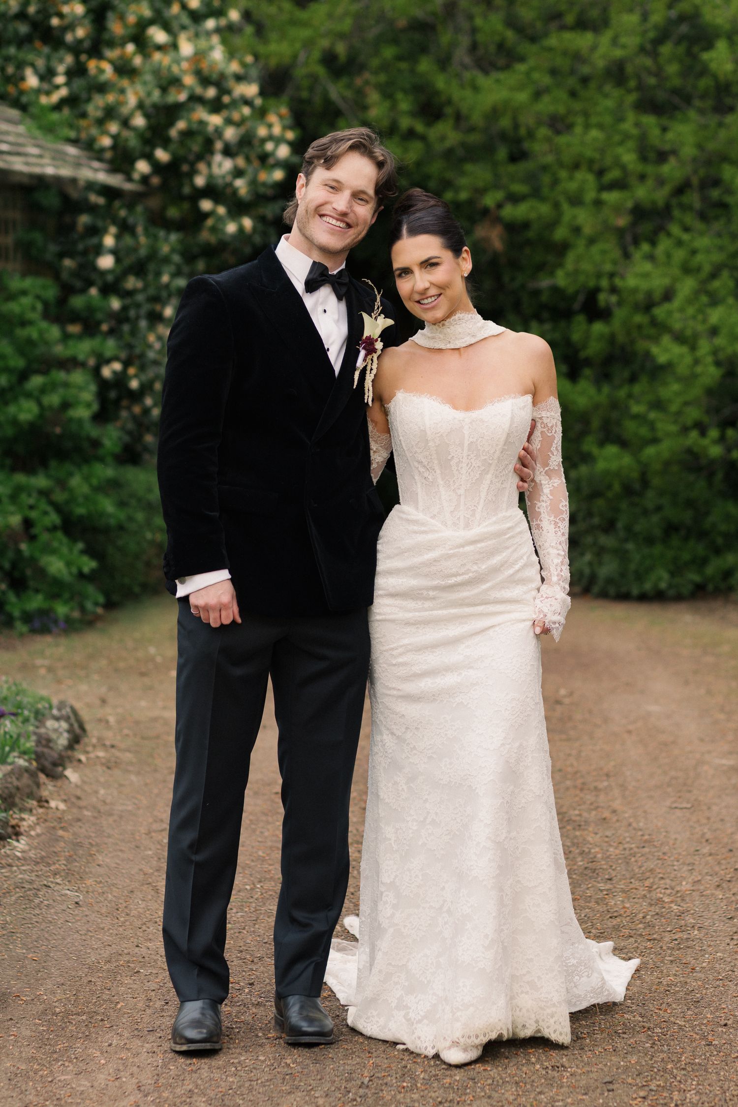 Smiling bride and groom posing together on a garden path, groom in black velvet tuxedo, bride in lace gown.