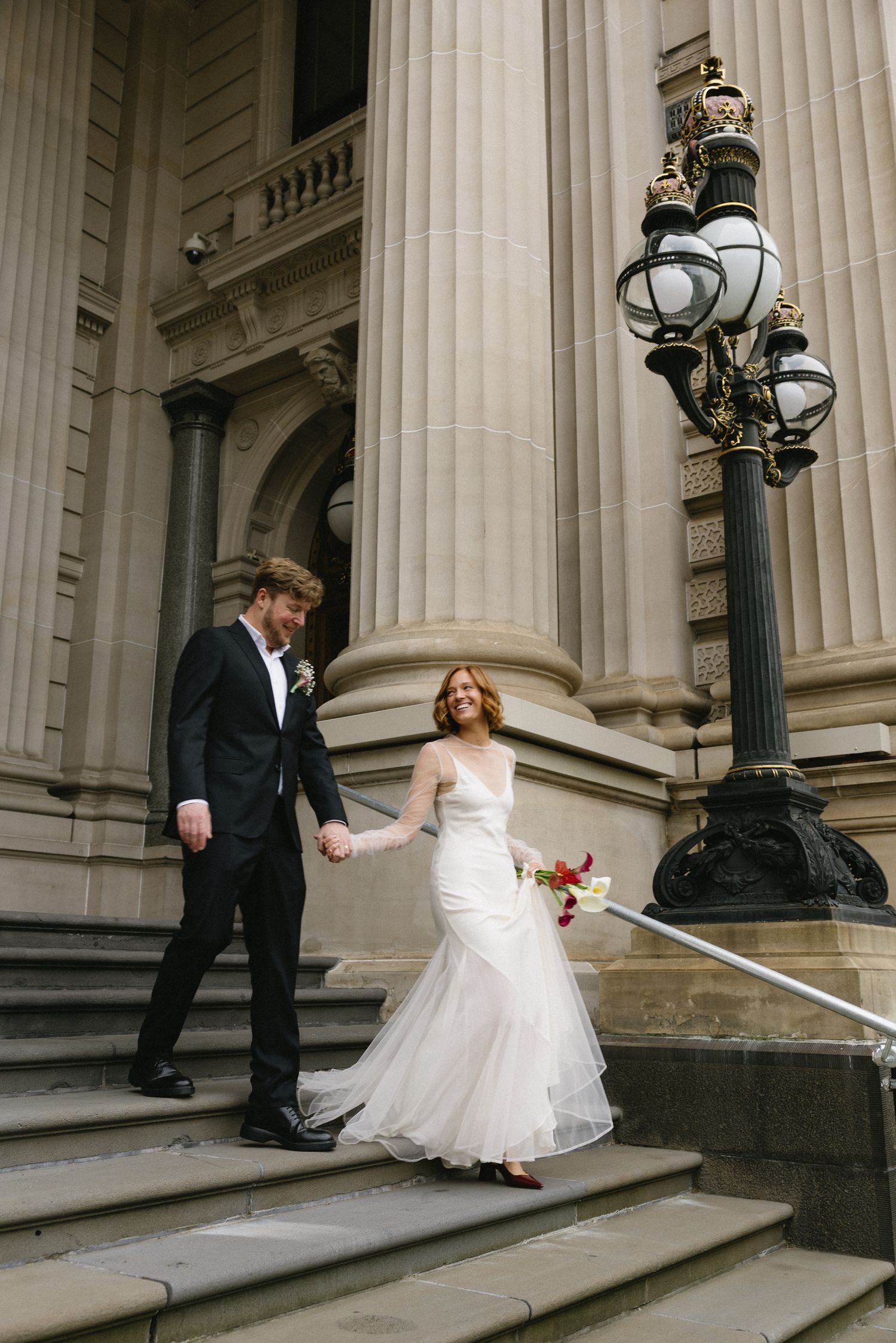 Bride and groom hold hands walking down stone steps of grand building with ornate lamp post.