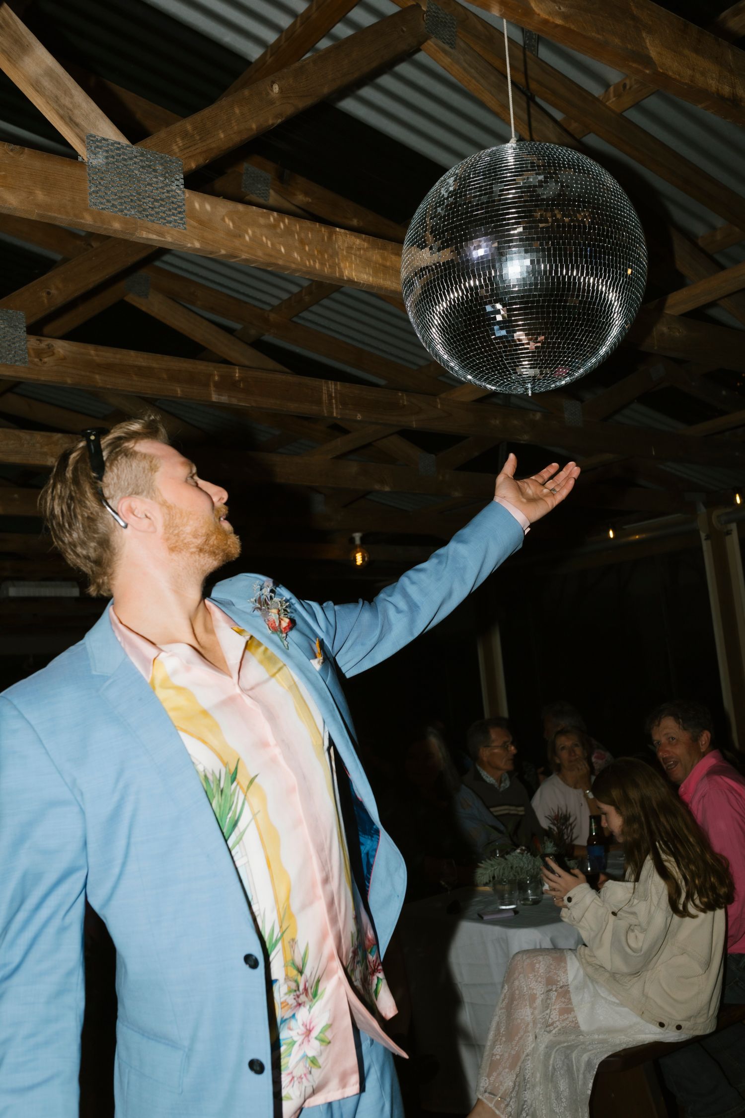 Man in a blue suit reaching up to touch a disco ball in a rustic barn wedding venue.