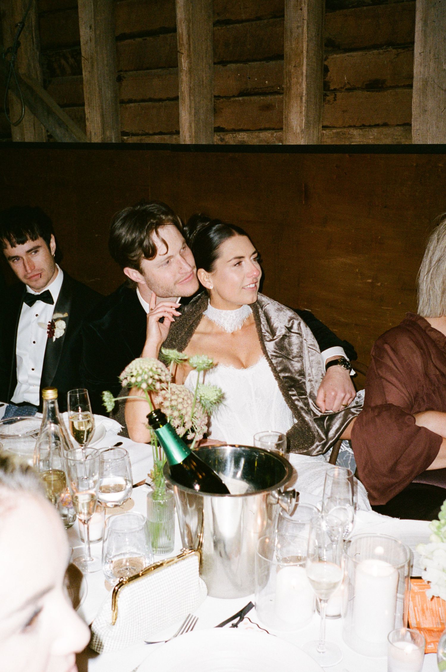 Bride and groom share an intimate moment at wedding reception table in rustic barn venue with champagne and floral centerpieces.