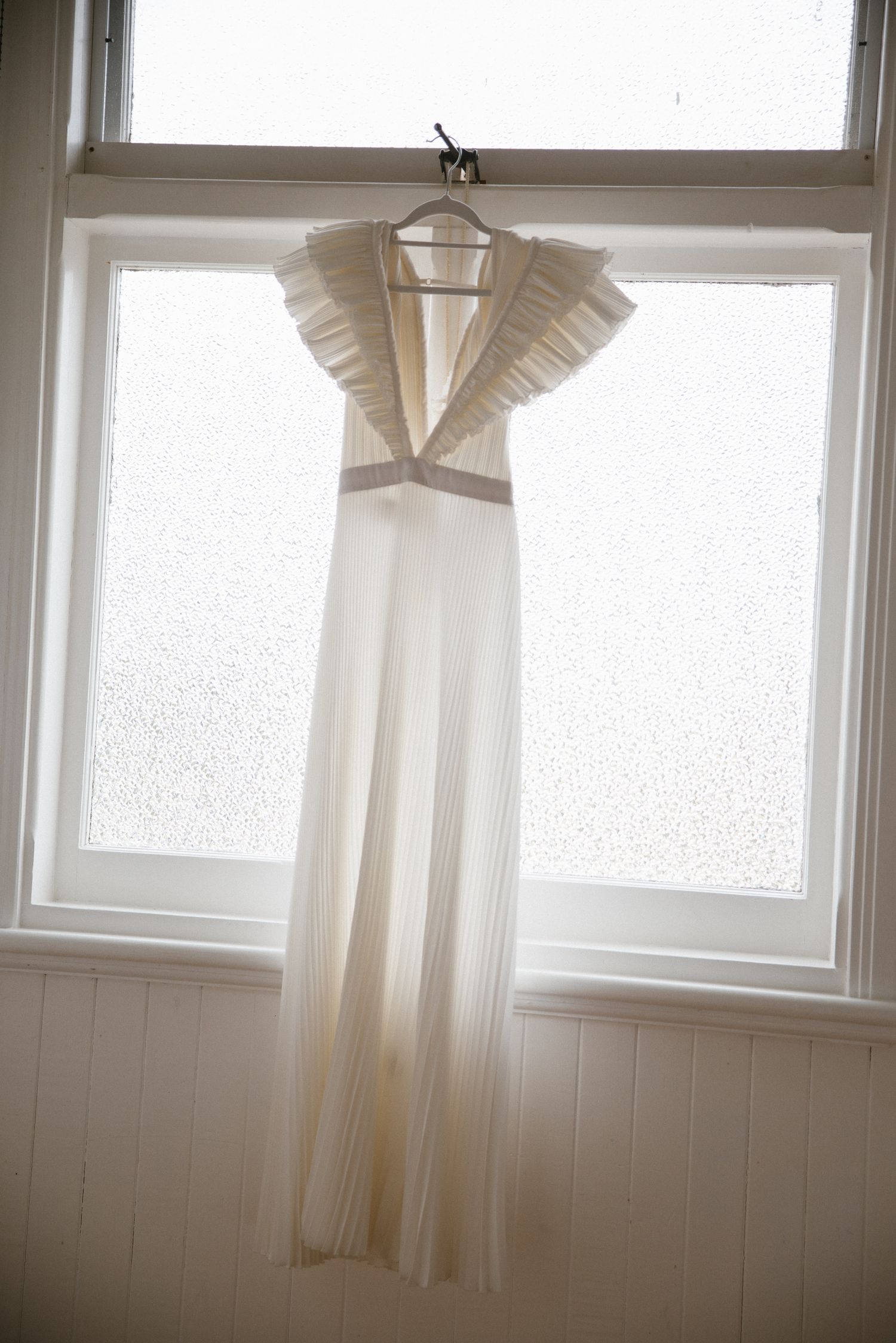 Elegant white pleated wedding dress with ruffled shoulders hanging in a sunlit window.
