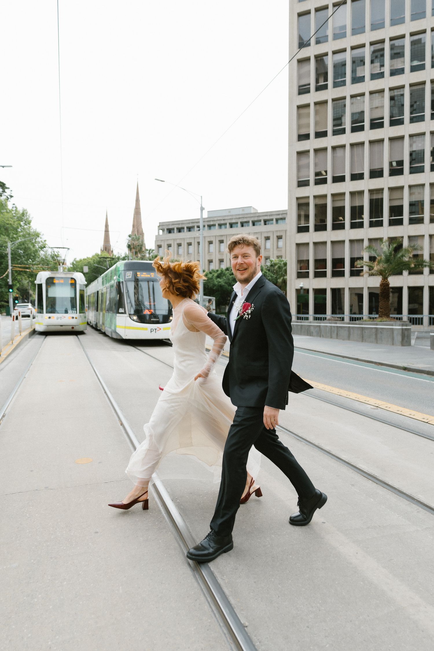 Bride and groom cross tram tracks in Melbourne CBD, modern trams and church spire visible in background.