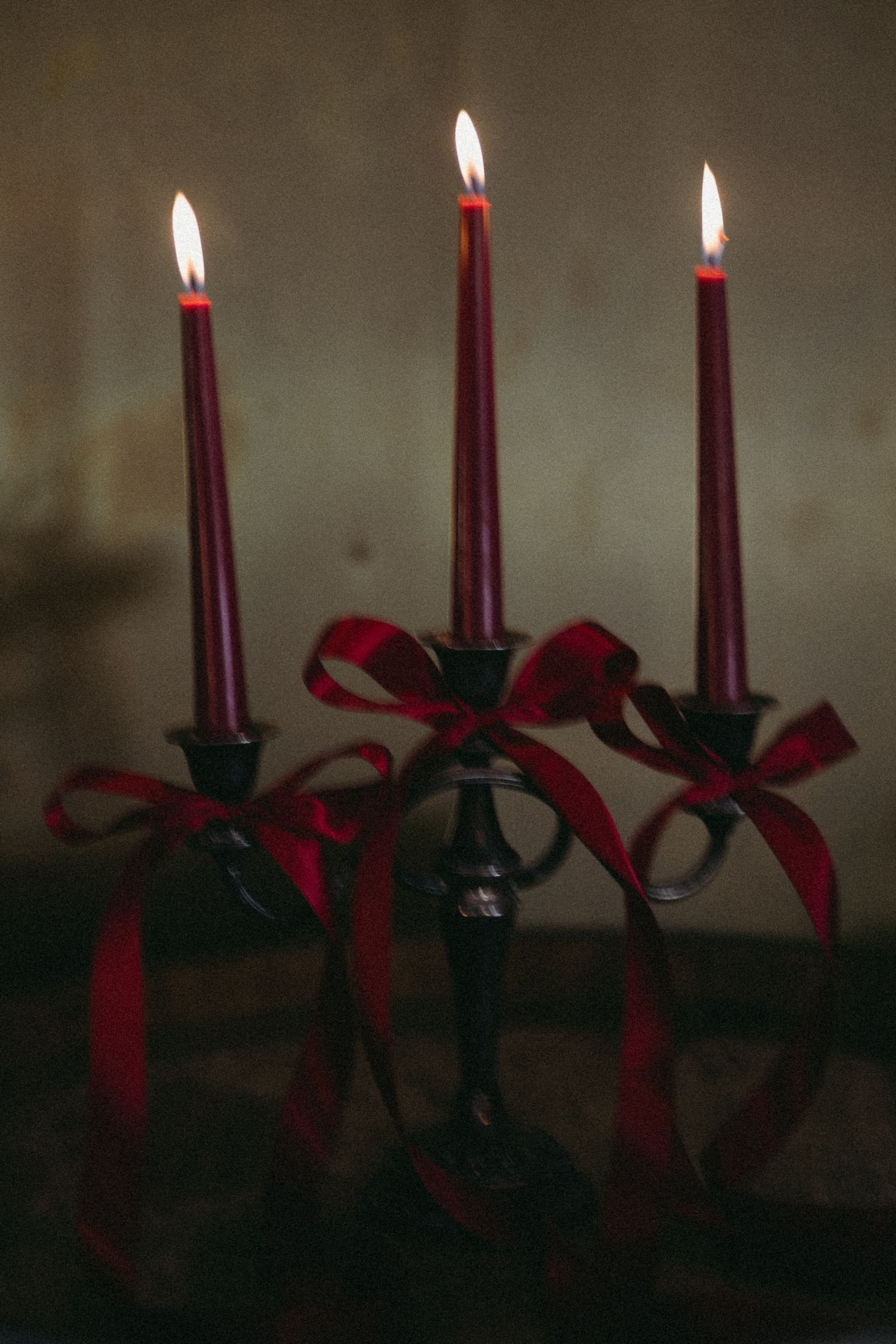 Three lit burgundy taper candles with red ribbon bows on black candelabra in moody setting.