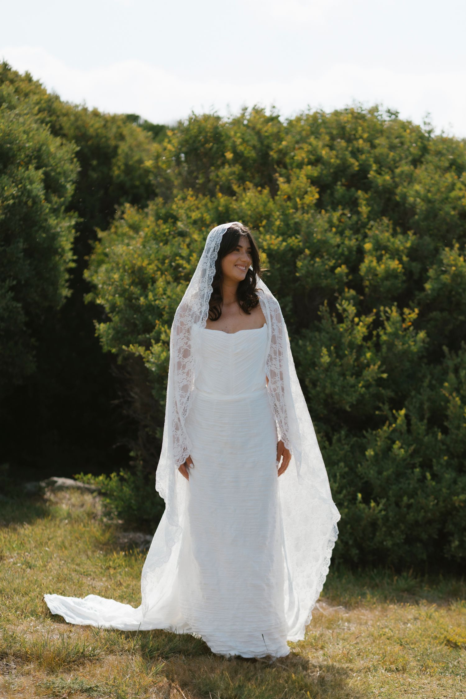 Bride in white lace gown and cathedral veil stands outdoors among green bushes on a sunny day.