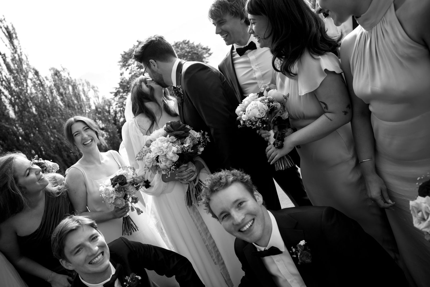 Black and white wedding party photo with bride and groom kissing surrounded by smiling bridesmaids and groomsmen outdoors.
