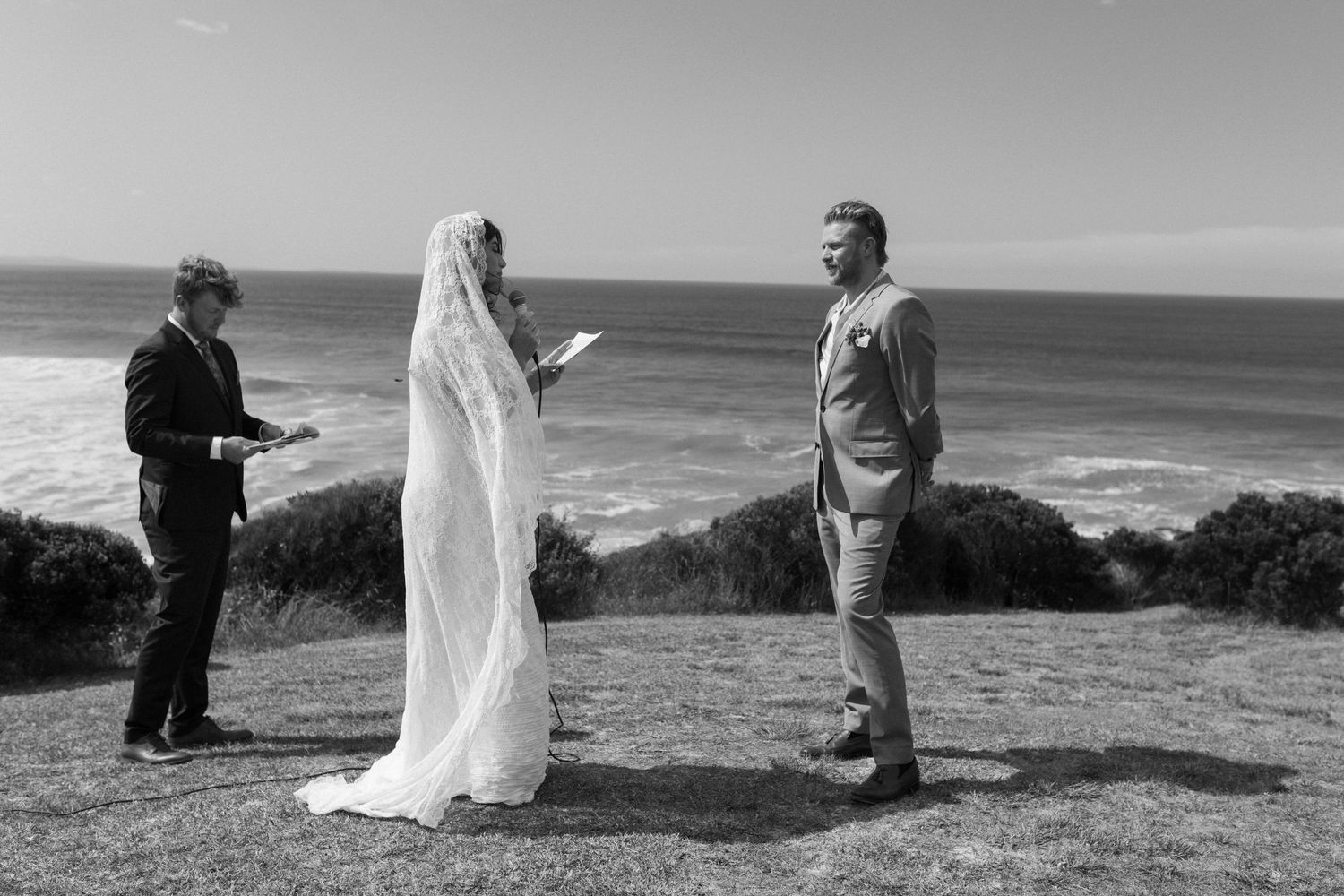 Black and white outdoor wedding ceremony by the ocean with bride, groom, and officiant on a cliff.