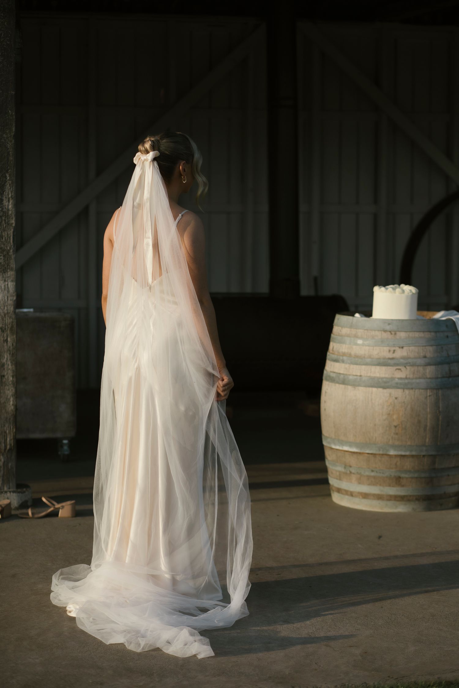Bride in white wedding dress and long veil standing in rustic barn with wooden barrel.