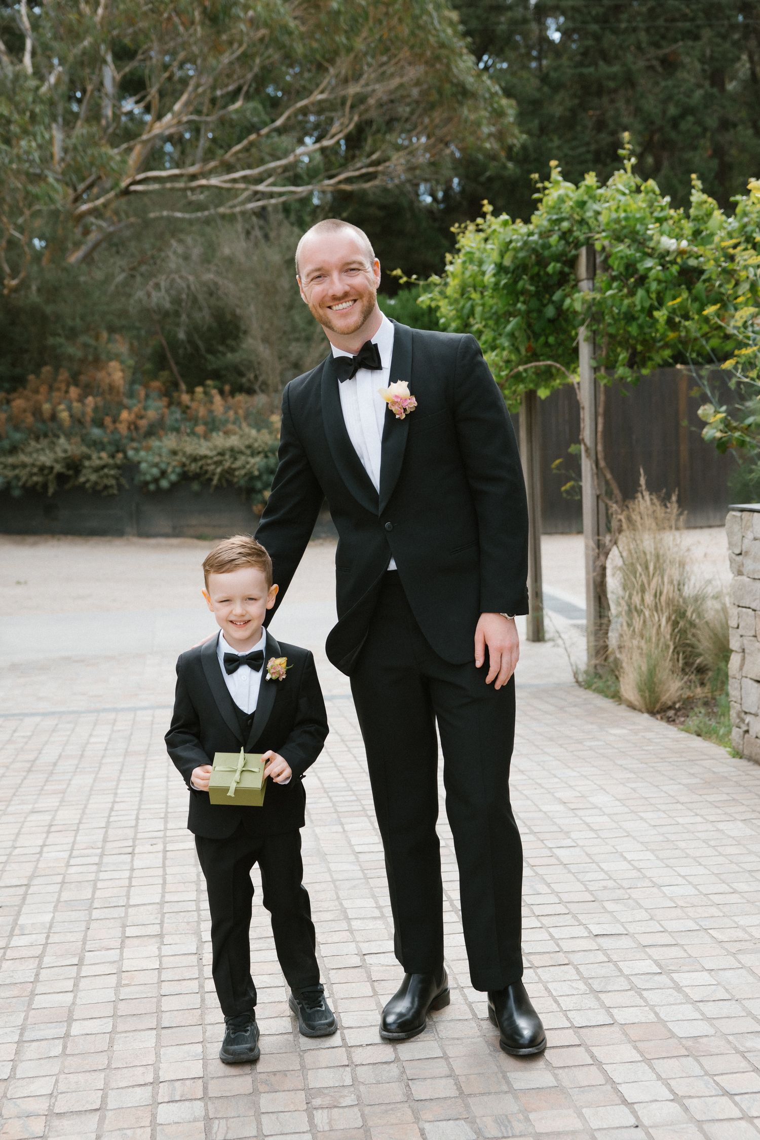 Smiling groom in black tuxedo standing with young ring bearer in matching suit holding gift box.