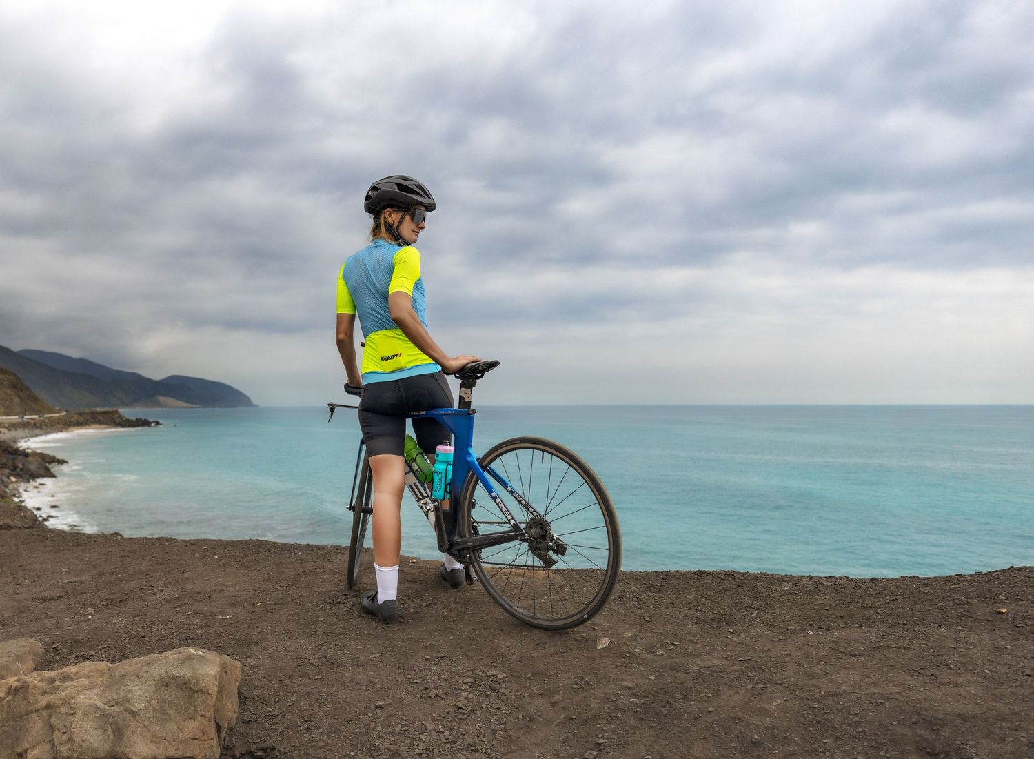 Female cyclist in yellow and blue kit standing with road bike overlooking a turquoise coastline.