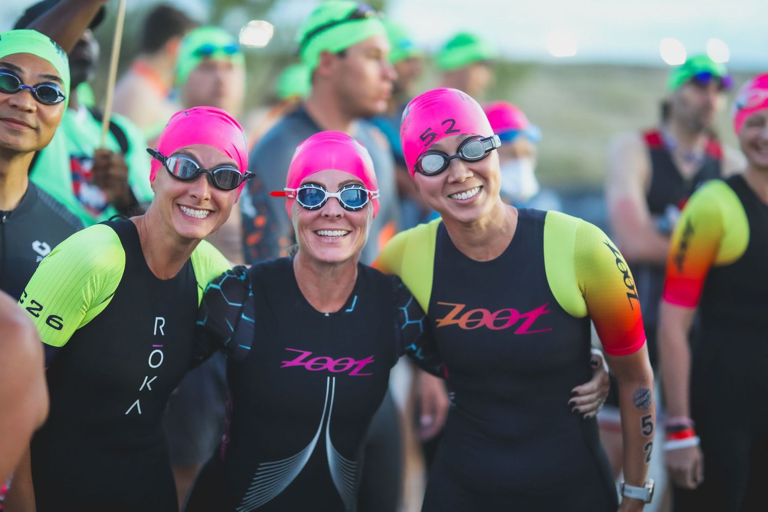 Three women in pink swim caps and wetsuits smile together at triathlon swim start.