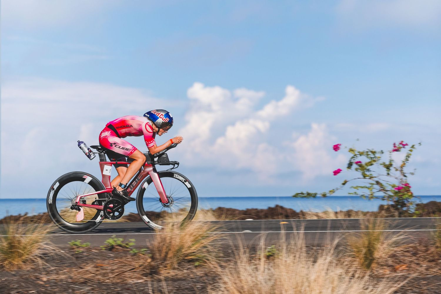 Cyclist in red kit racing on triathlon bike along scenic coastal highway.