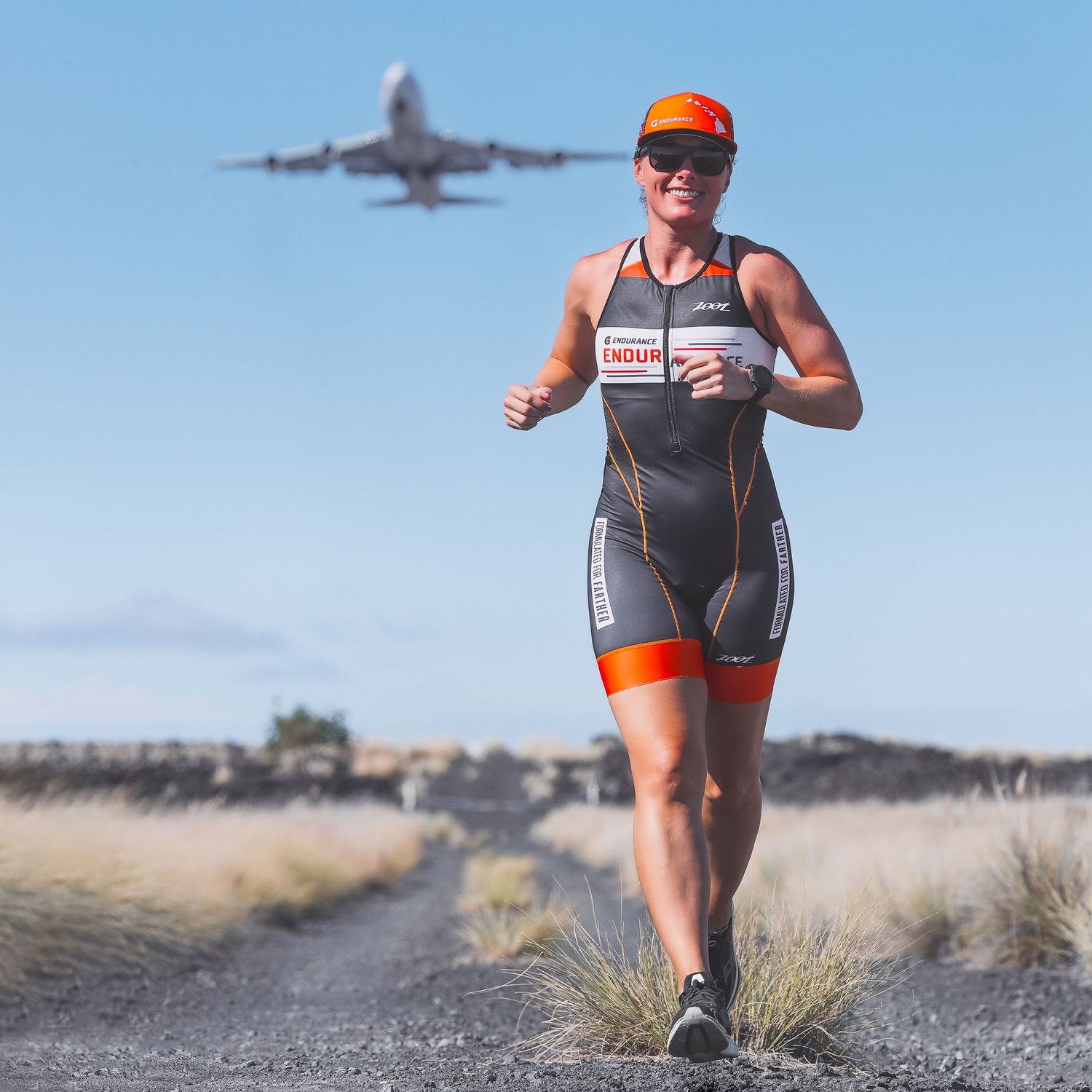 Female triathlete running in race suit with airplane flying overhead in background.