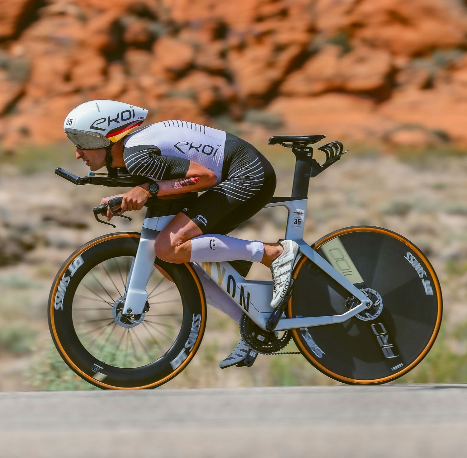 Triathlete in EKOI black kit rides a time trial bike in an aggressive aero position past red rock formations.