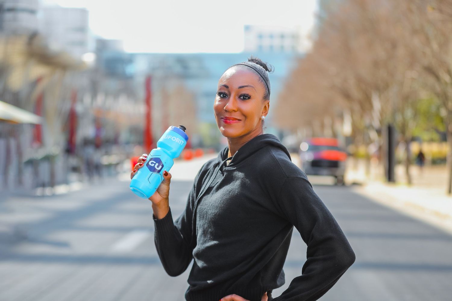 Smiling woman in black hoodie holding a GU energy drink bottle on a tree-lined urban path.