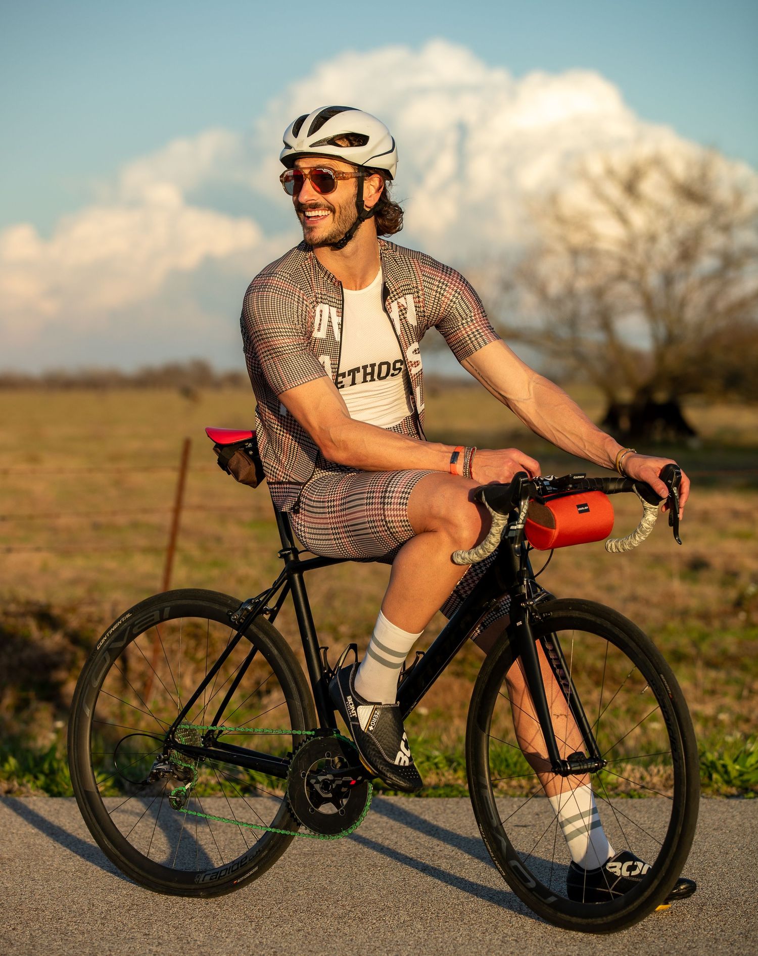 Smiling male cyclist riding black road bike in plaid kit with white helmet outdoors.