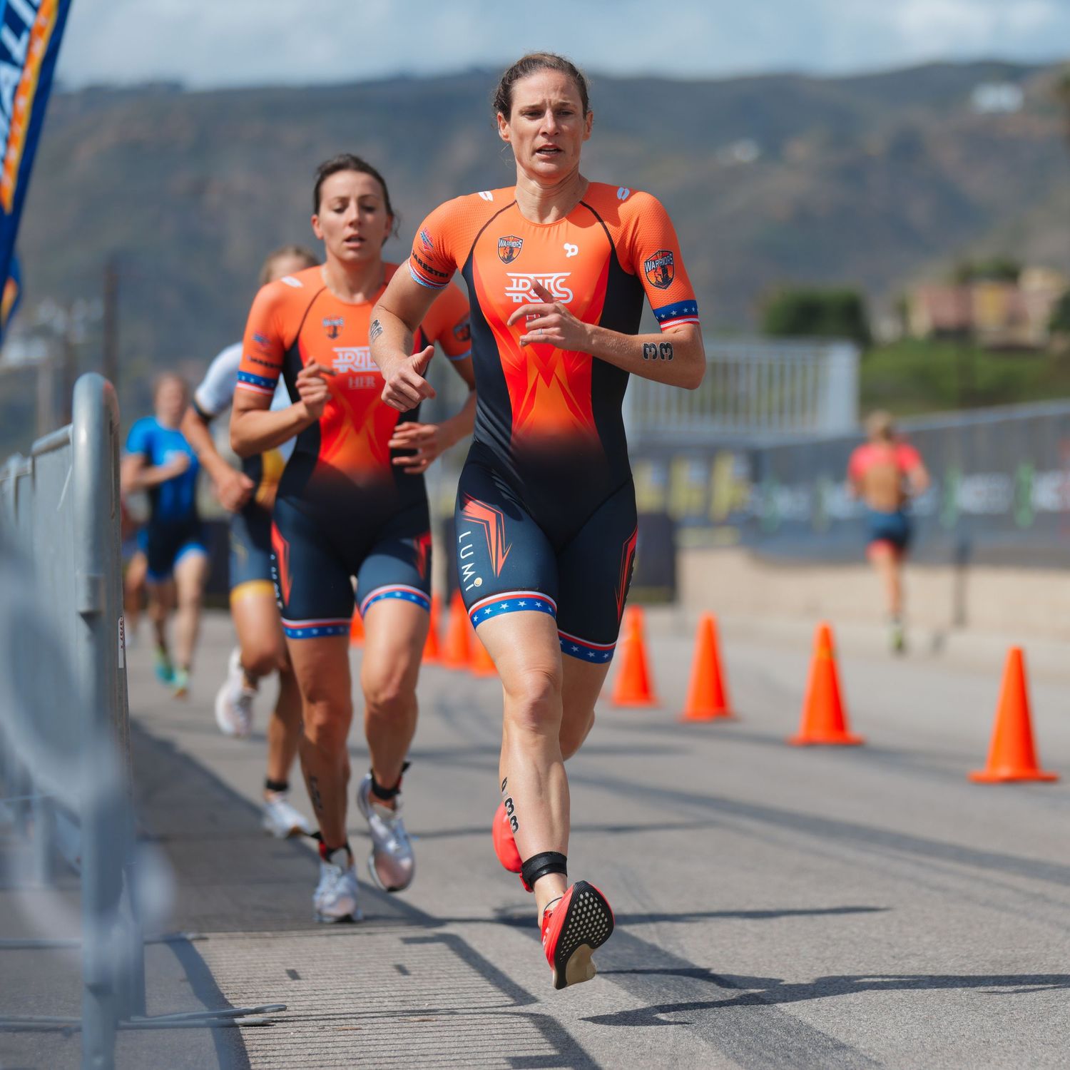 Female triathletes in orange USA kits run competitively along a coned course during a triathlon event.