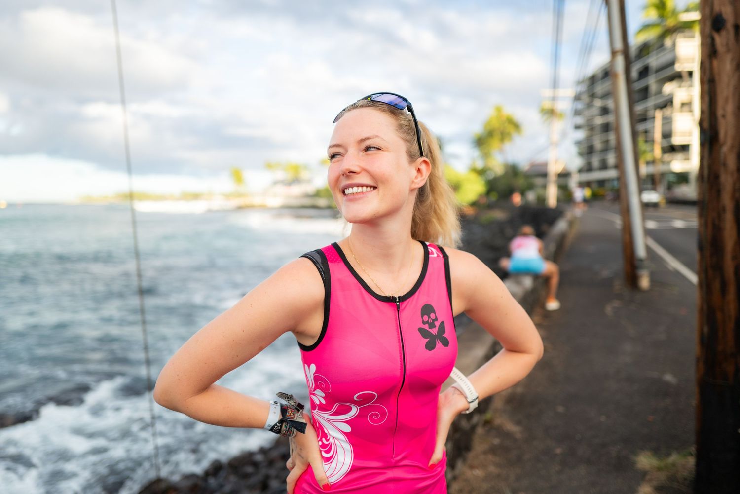 Smiling female athlete in pink triathlon suit stands near rocky Hawaiian coastline with palm trees.