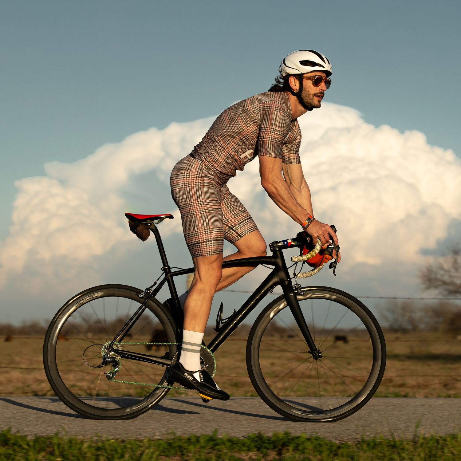 Male cyclist riding a road bike wearing a plaid cycling kit and white helmet with clouds in background.