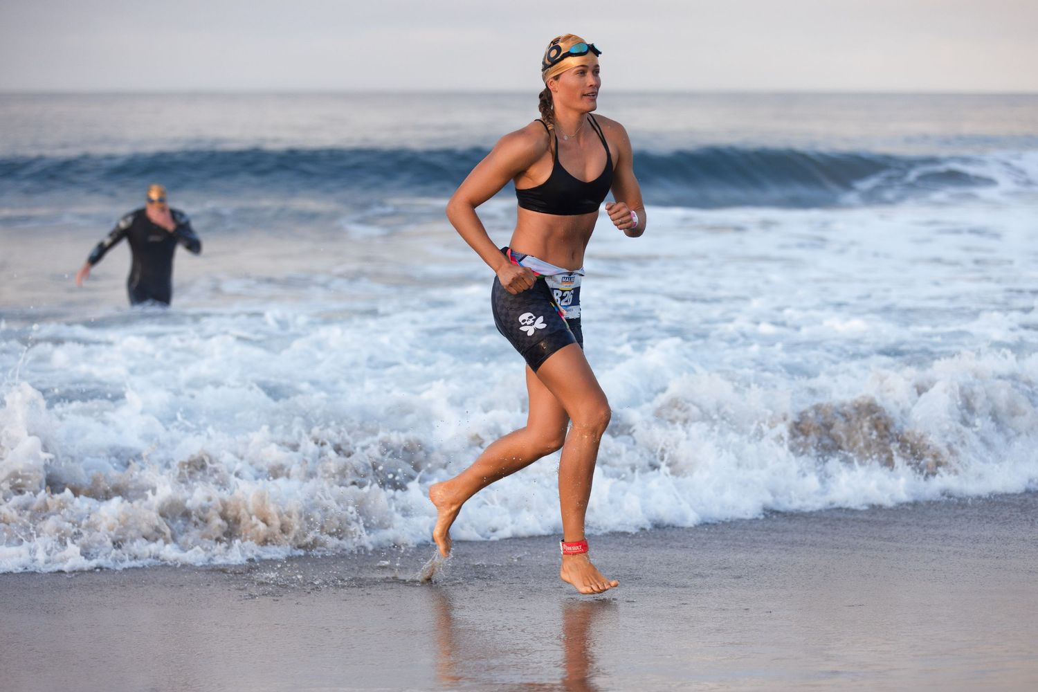 Female triathlete running barefoot along ocean shoreline wearing black sports bra and shorts after swim leg.