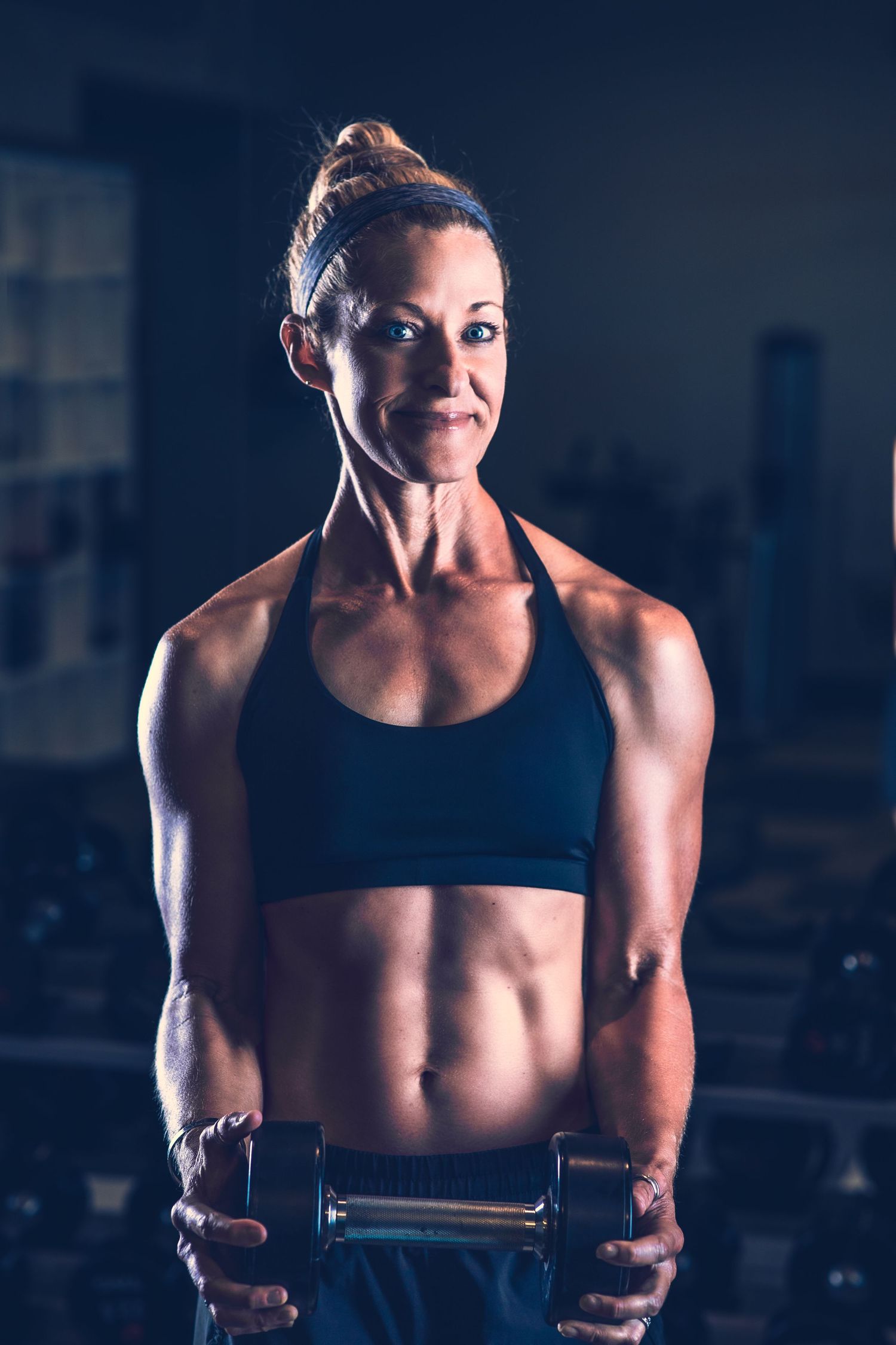 Muscular woman in black sports bra holding dumbbells in dramatic gym lighting.