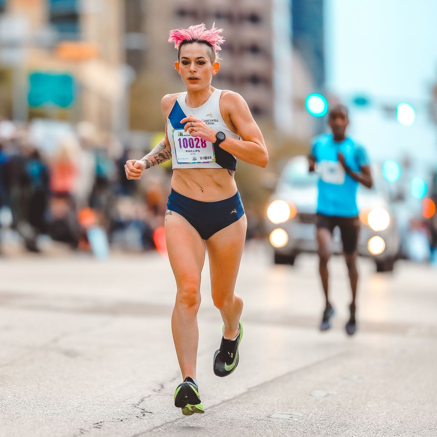 Female runner with pink hair wearing race bib 10028 competes in urban marathon race on city street.