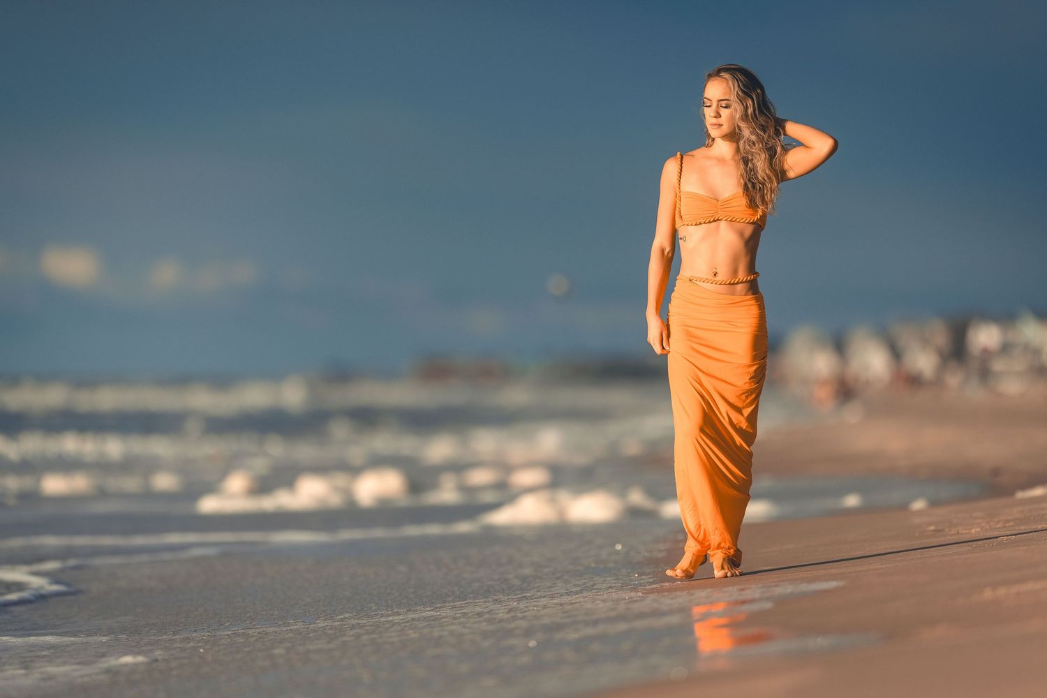 Blonde woman in orange satin crop top and maxi skirt walking along beach at sunset.