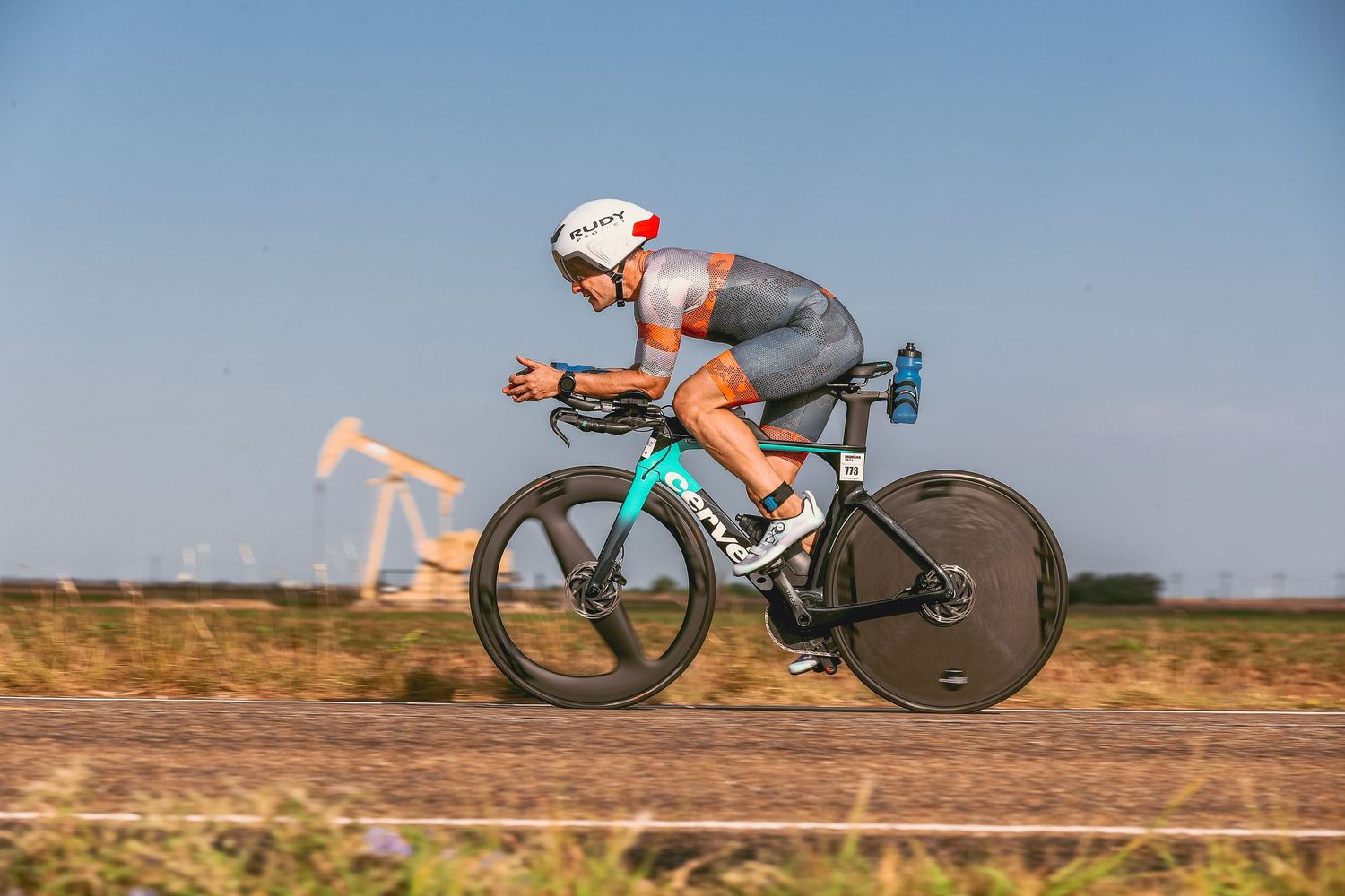 Cyclist in aerodynamic position races on a time trial bike through flat open countryside terrain.