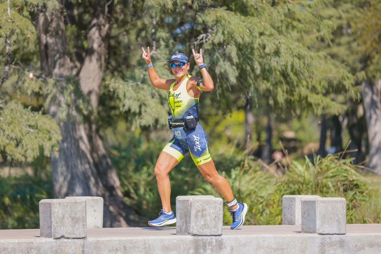 Triathlete running outdoors giving peace signs wearing yellow racing kit and blue shoes during a sunny race event.