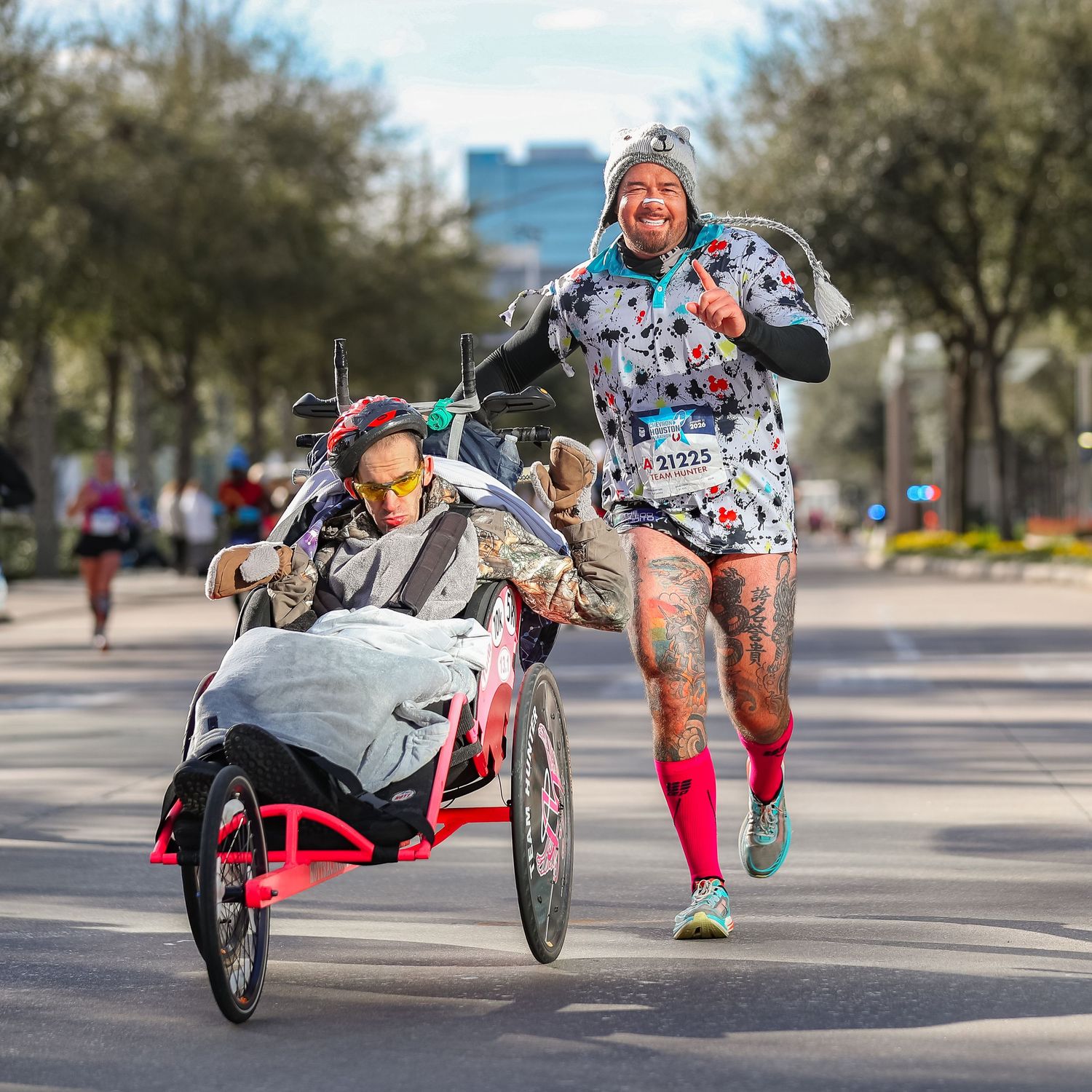 Tattooed runner in festive costume pushes a participant in a racing wheelchair during a road race.
