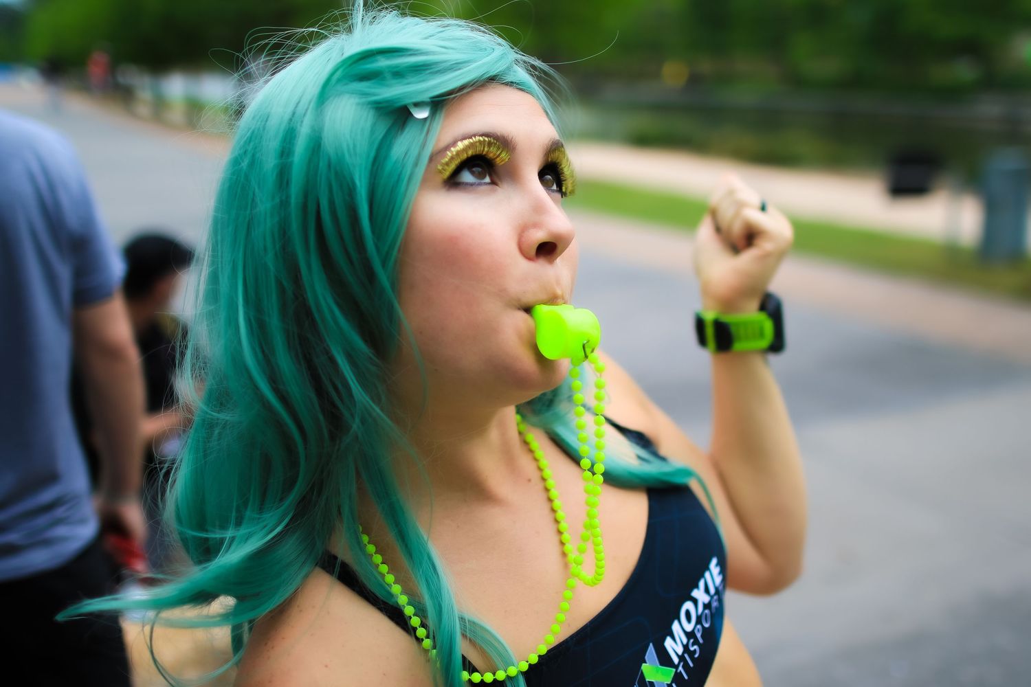 Woman with teal hair blowing green whistle wearing Moxie tank top at race event.