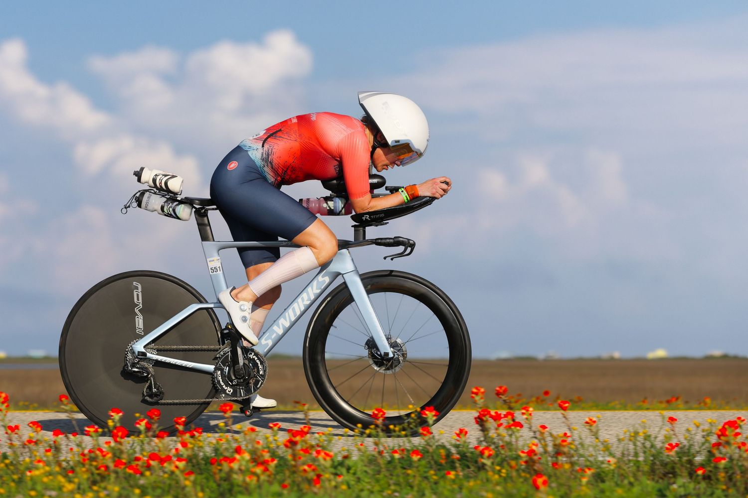 Cyclist in red kit rides aerodynamic time trial bike past red poppies under blue sky.