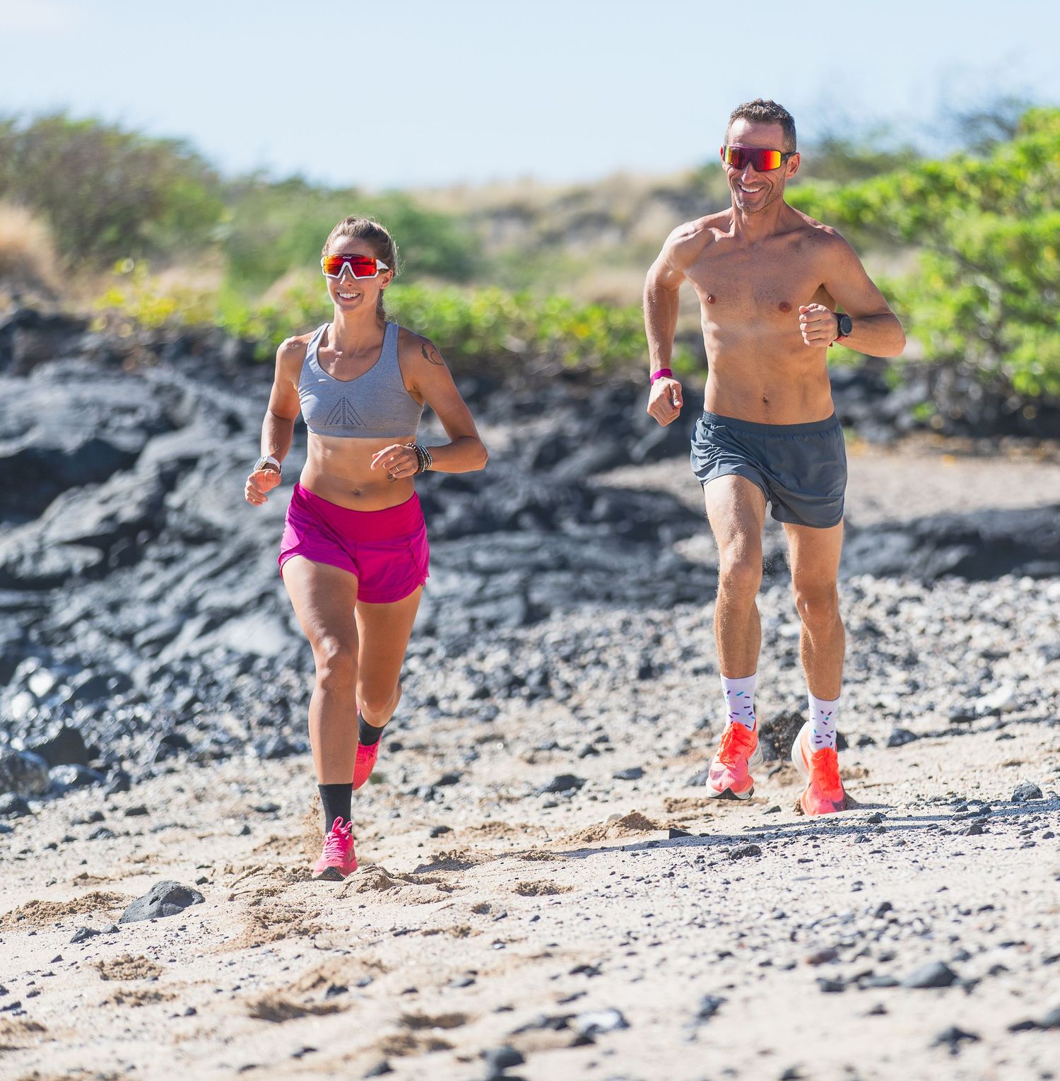 Man and woman running together on rocky beach trail in sunglasses.