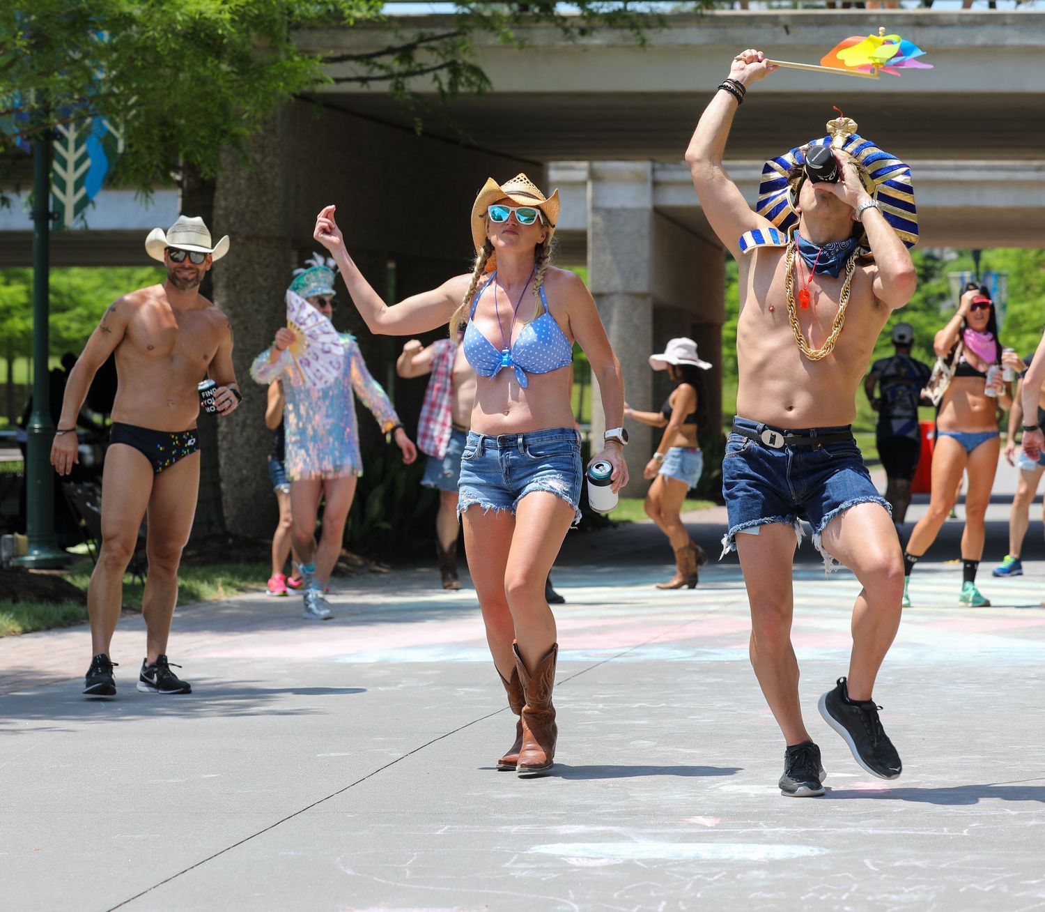 People dancing outdoors in summer festival attire including bikinis, cowboy hats, and denim shorts on a sunny day.