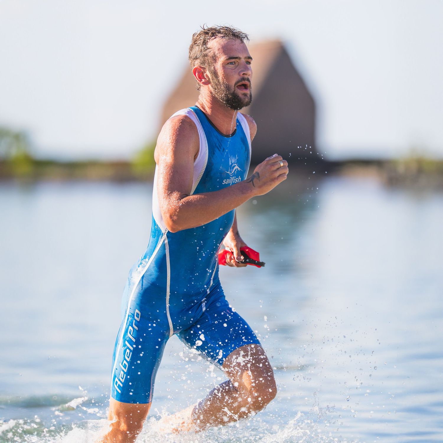 Male triathlete in blue racing suit splashes through shallow water during a triathlon transition.
