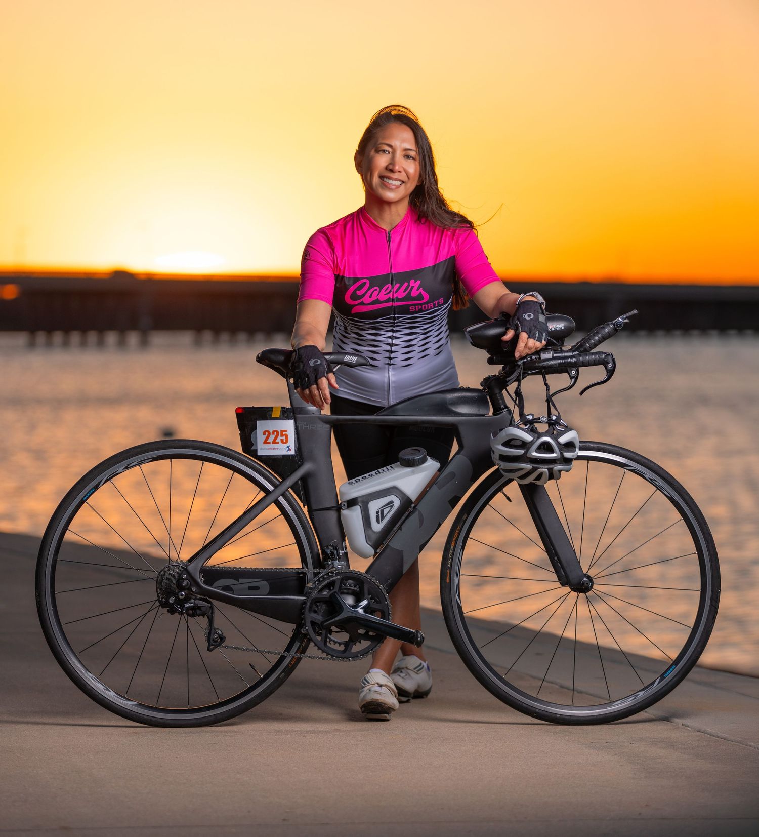Woman in pink Coeur cycling jersey posing with triathlon bike at sunset near waterfront at golden hour.