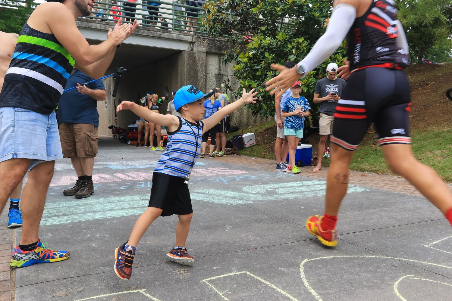 Young boy in blue cap high-fiving runners during outdoor triathlon race event.