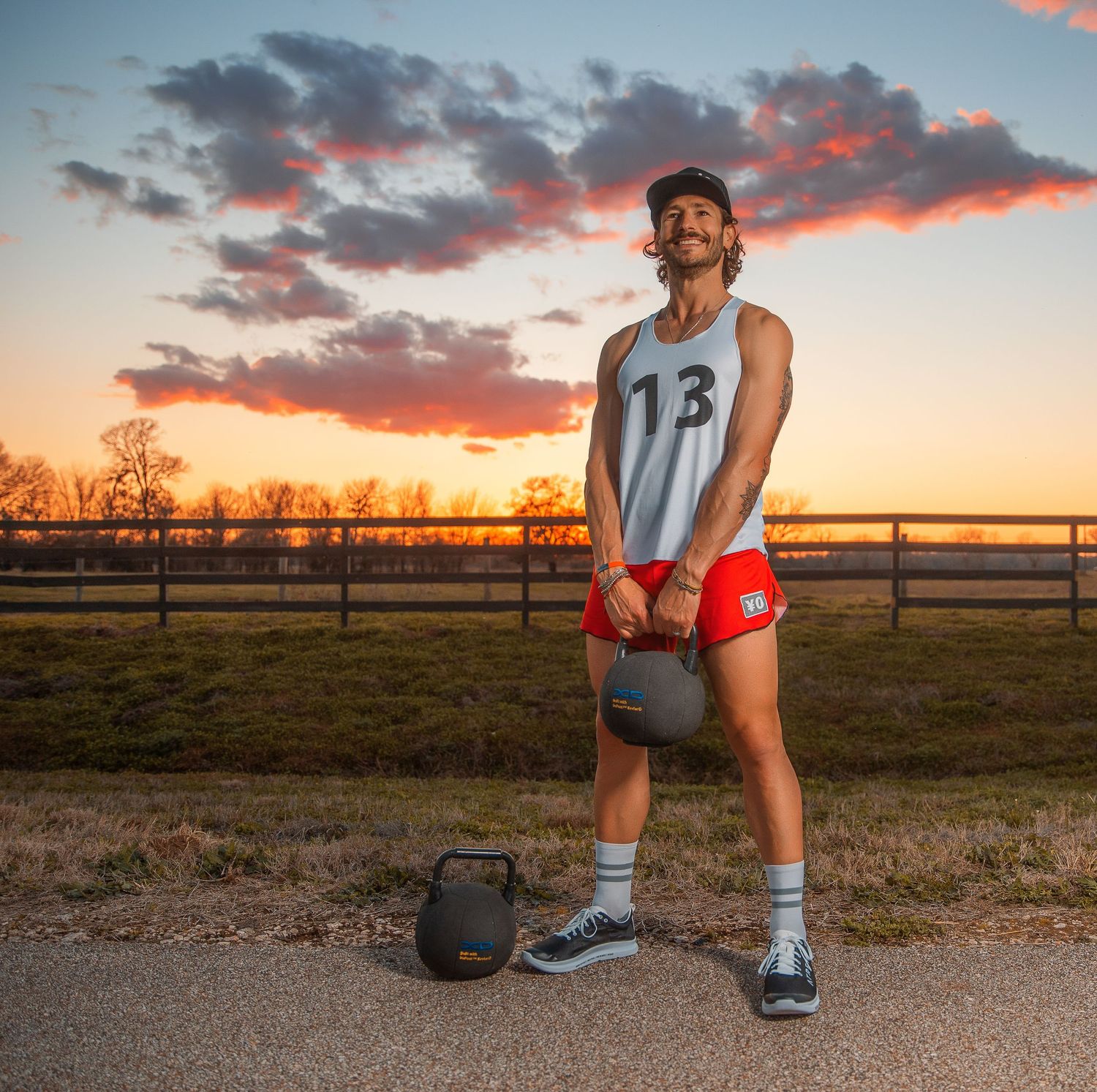 Athletic man in number 13 tank top holds kettlebell on rural path at sunset.