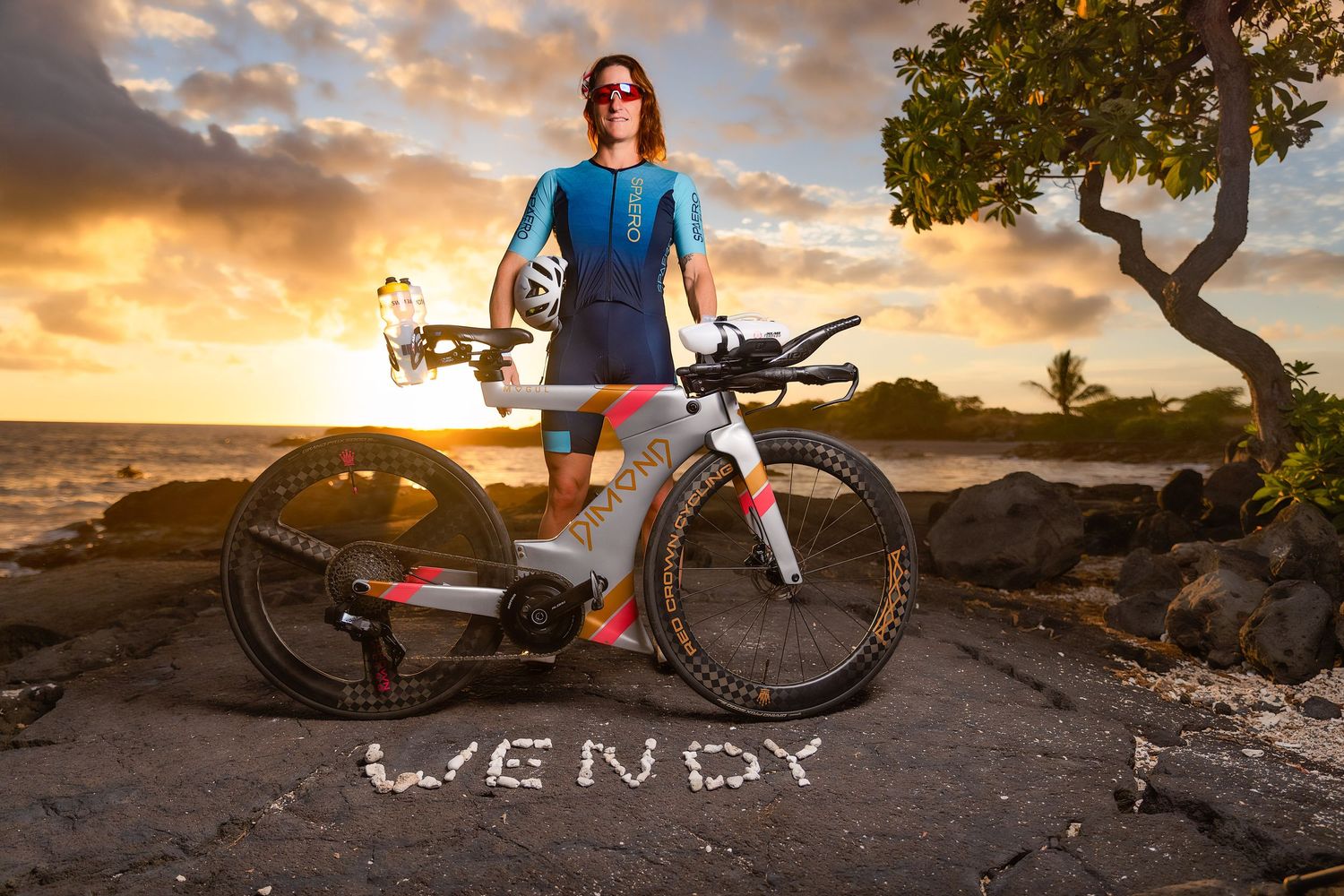 Female triathlete poses with bike at sunset, name WENDY written on ground.