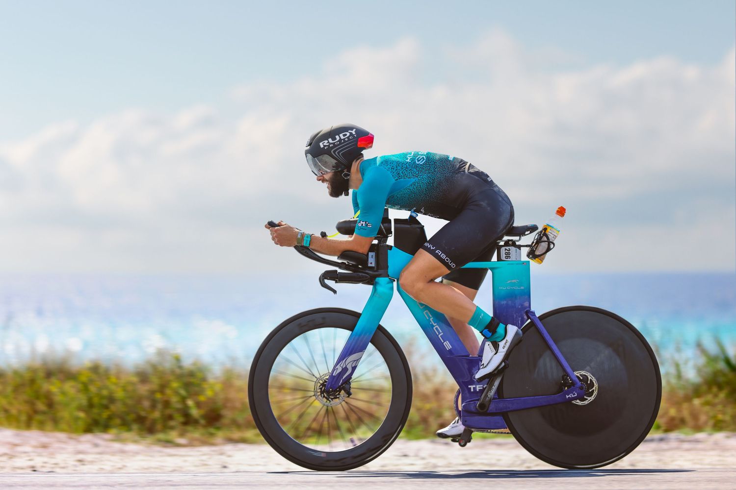 Triathlete in teal kit cycling aerodynamically along scenic beach road.