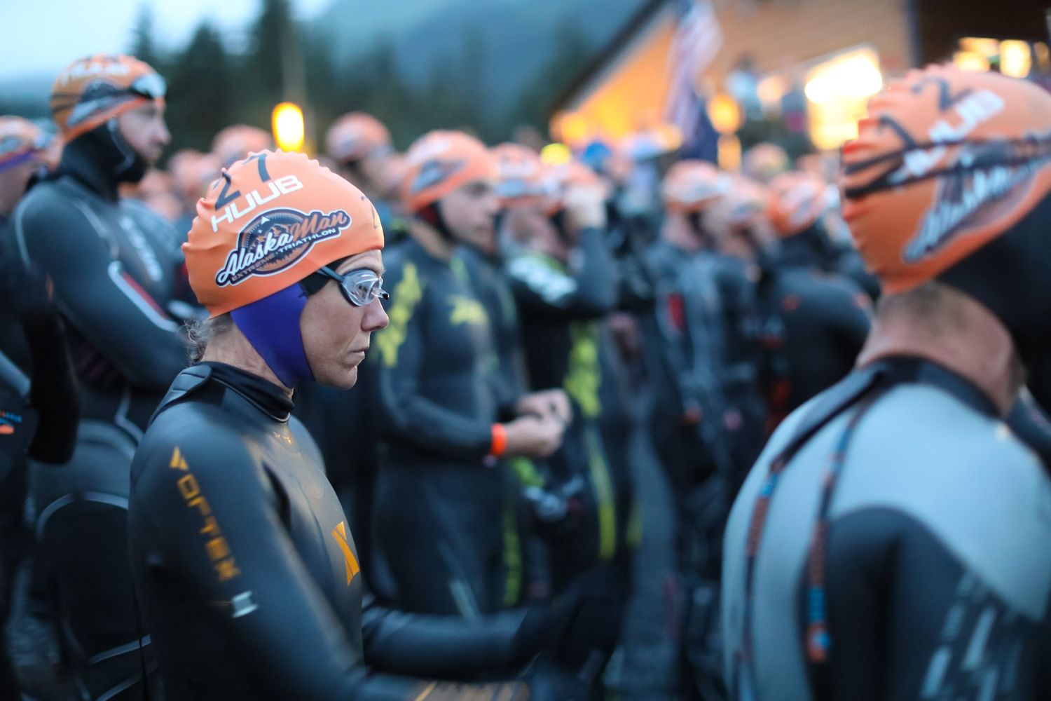 Triathletes in orange swim caps and wetsuits gather at dawn before an Ironman swim start.