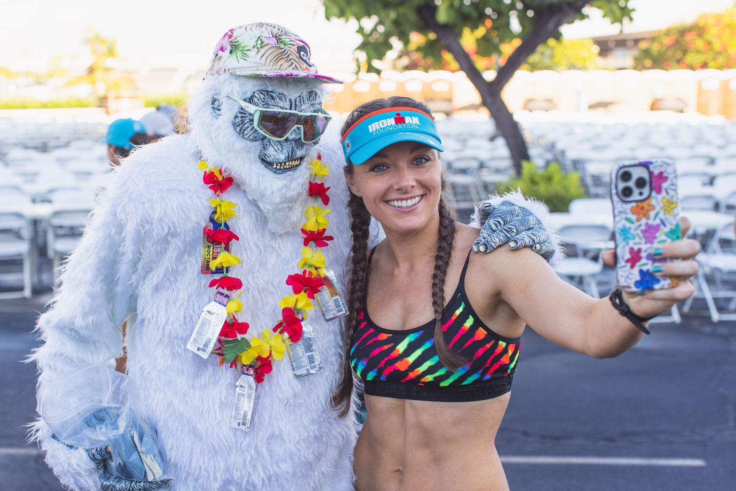 Female athlete takes selfie with person dressed in Yeti costume wearing Hawaiian lei at triathlon event.