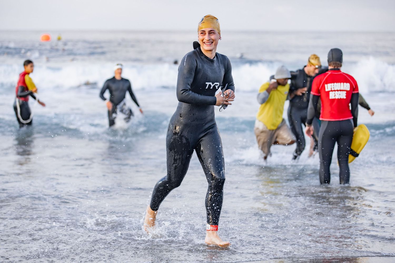 Smiling triathlete in wetsuit emerges from ocean waves during an open water swimming competition.
